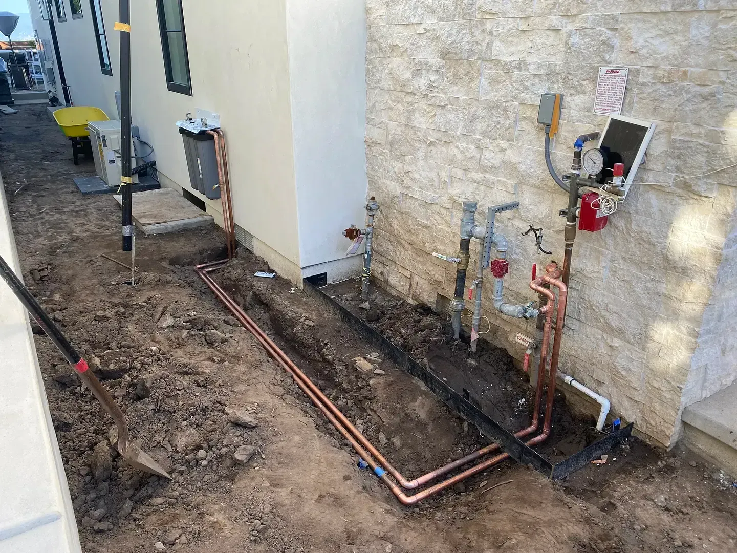 Construction site with exposed copper pipes, fittings, and gas meter against a stone wall. Dirt and tools are also present.