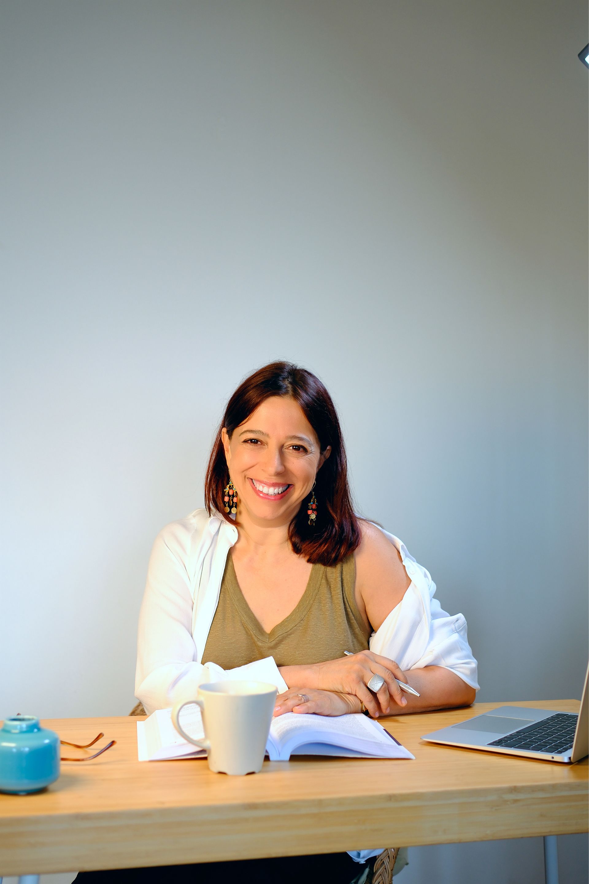 Mulher de cabelos escuros sorri em uma mesa, trabalhando em um laptop, com uma xícara de café e um bloco de notas.