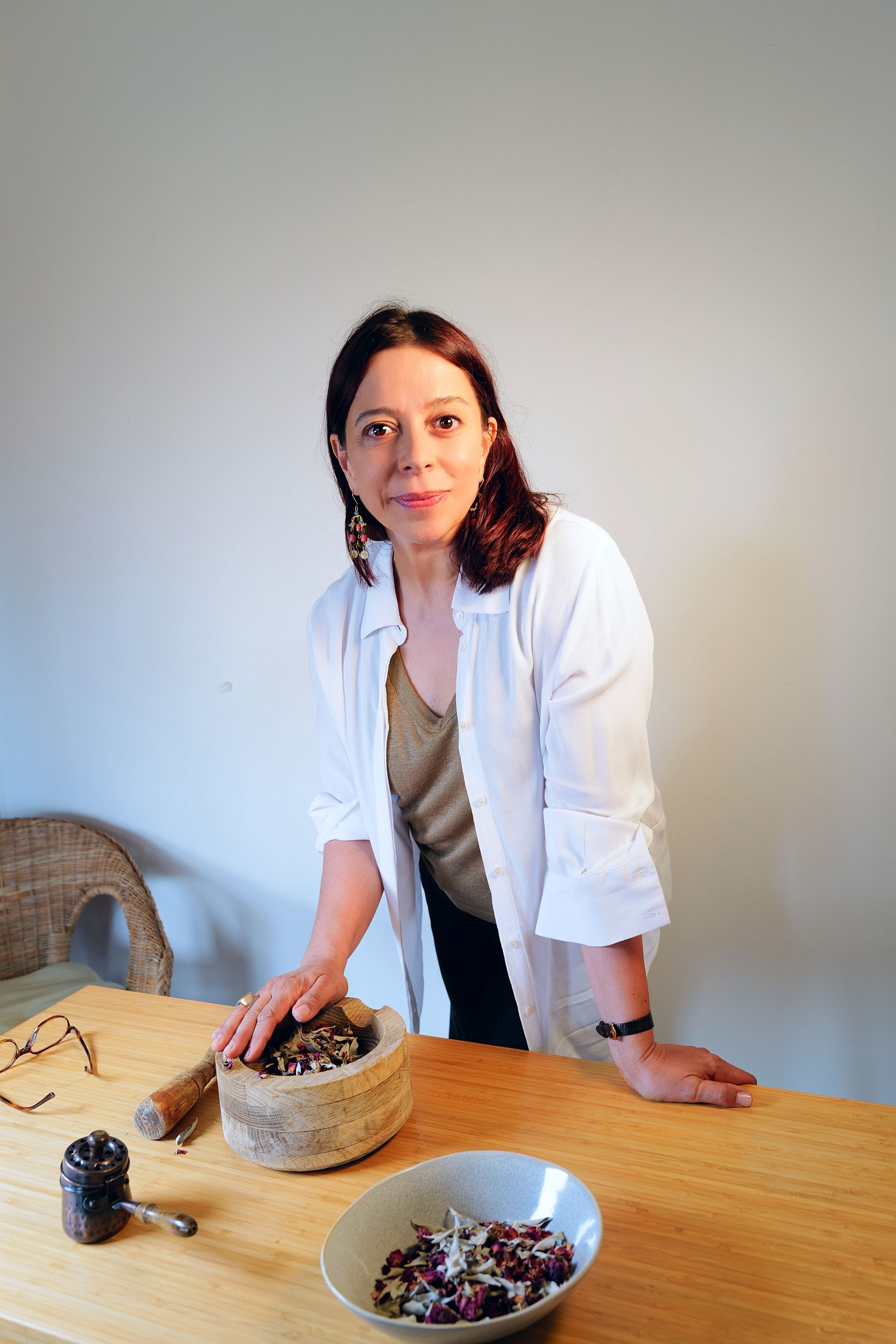 Dra Patricia Sofia Homeopatia ndebruçada sobre uma mesa com pilão e almofariz, e um vaso de flores, em uma sala.