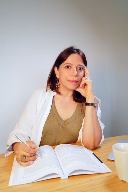 Mulher sentada a uma mesa de madeira, escrevendo em um livro aberto. Ela apoia o queixo na mão e parece pensativa.