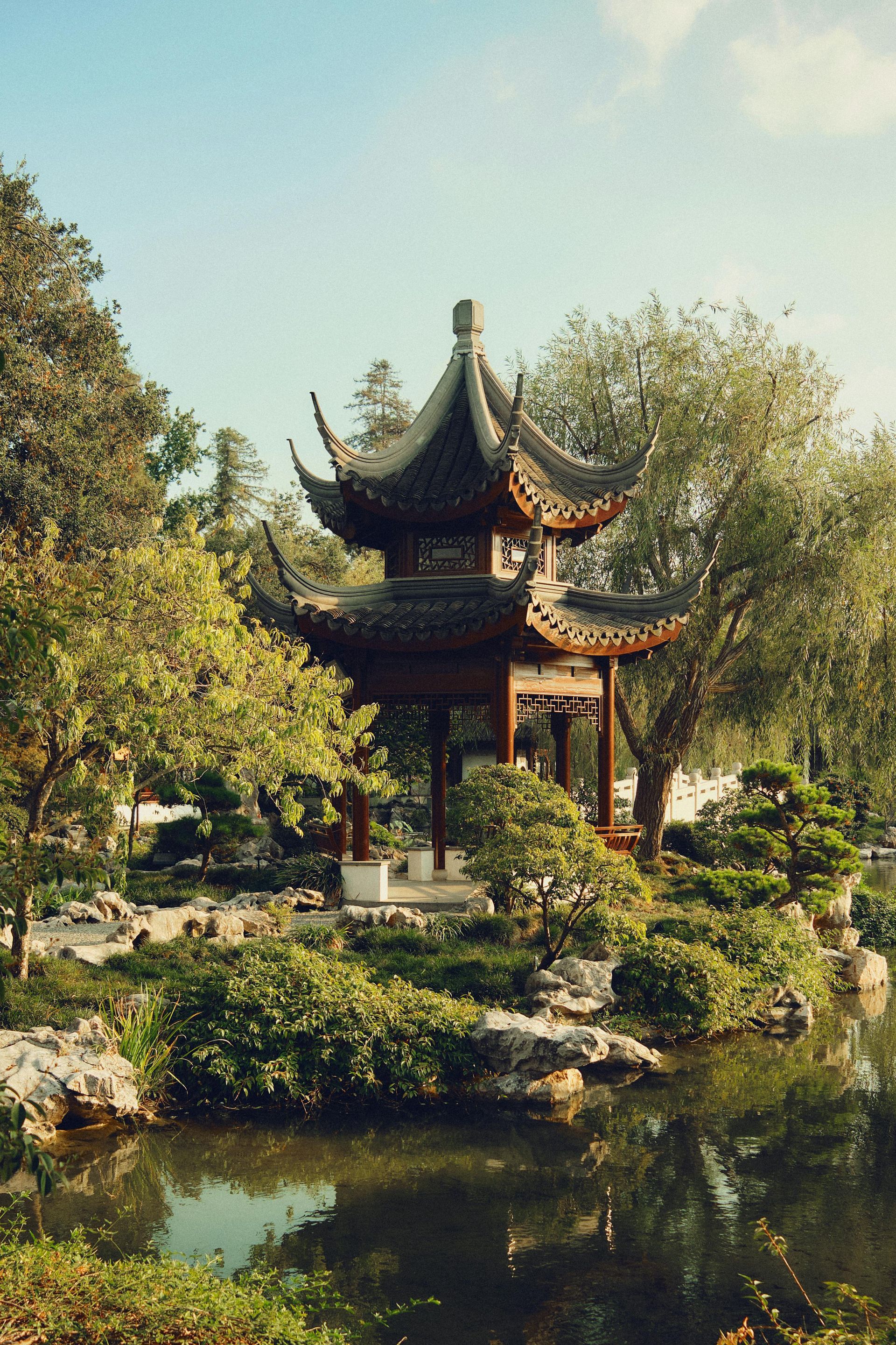 Um pagode chinês tradicional com teto escuro, refletido em um lago, cercado por árvores e vegetação exuberante.