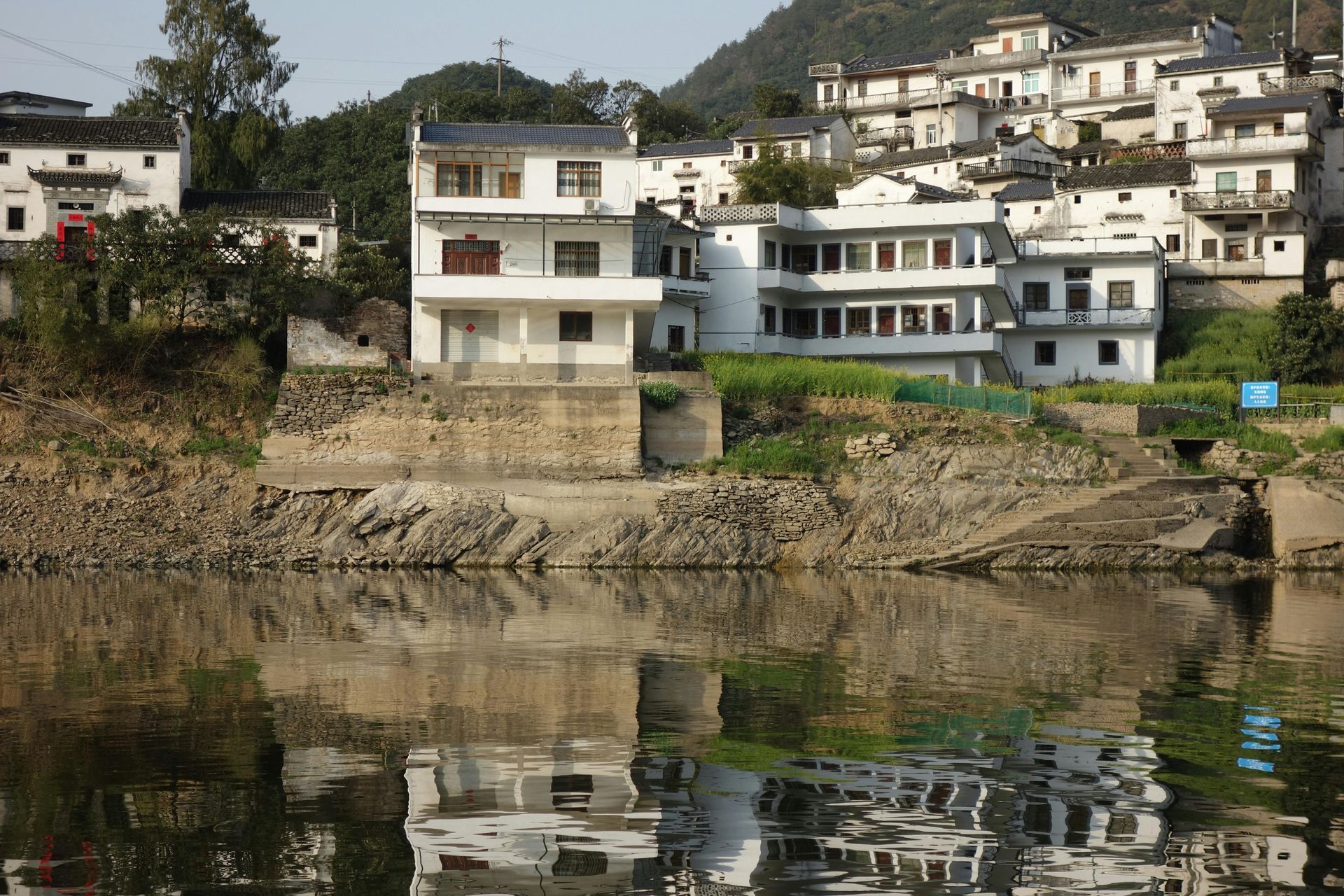 Prédios brancos com telhados escuros erguem-se de um barranco rochoso, refletindo-se nas ondas da água. 