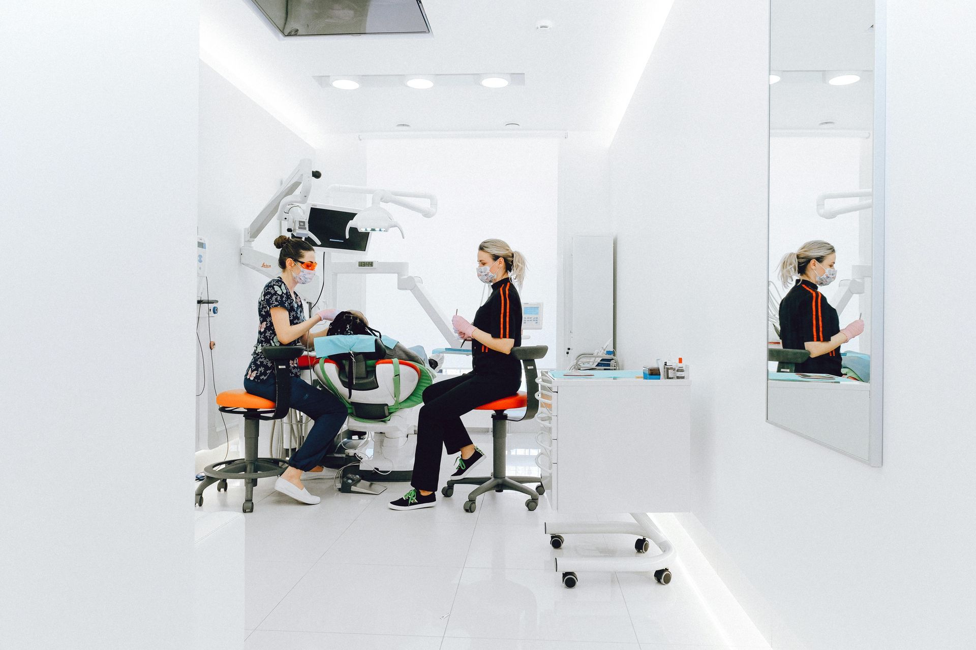 Duas mulheres em um consultório odontológico branco; uma em uma cadeira com ferramentas, a outra examinando um paciente.