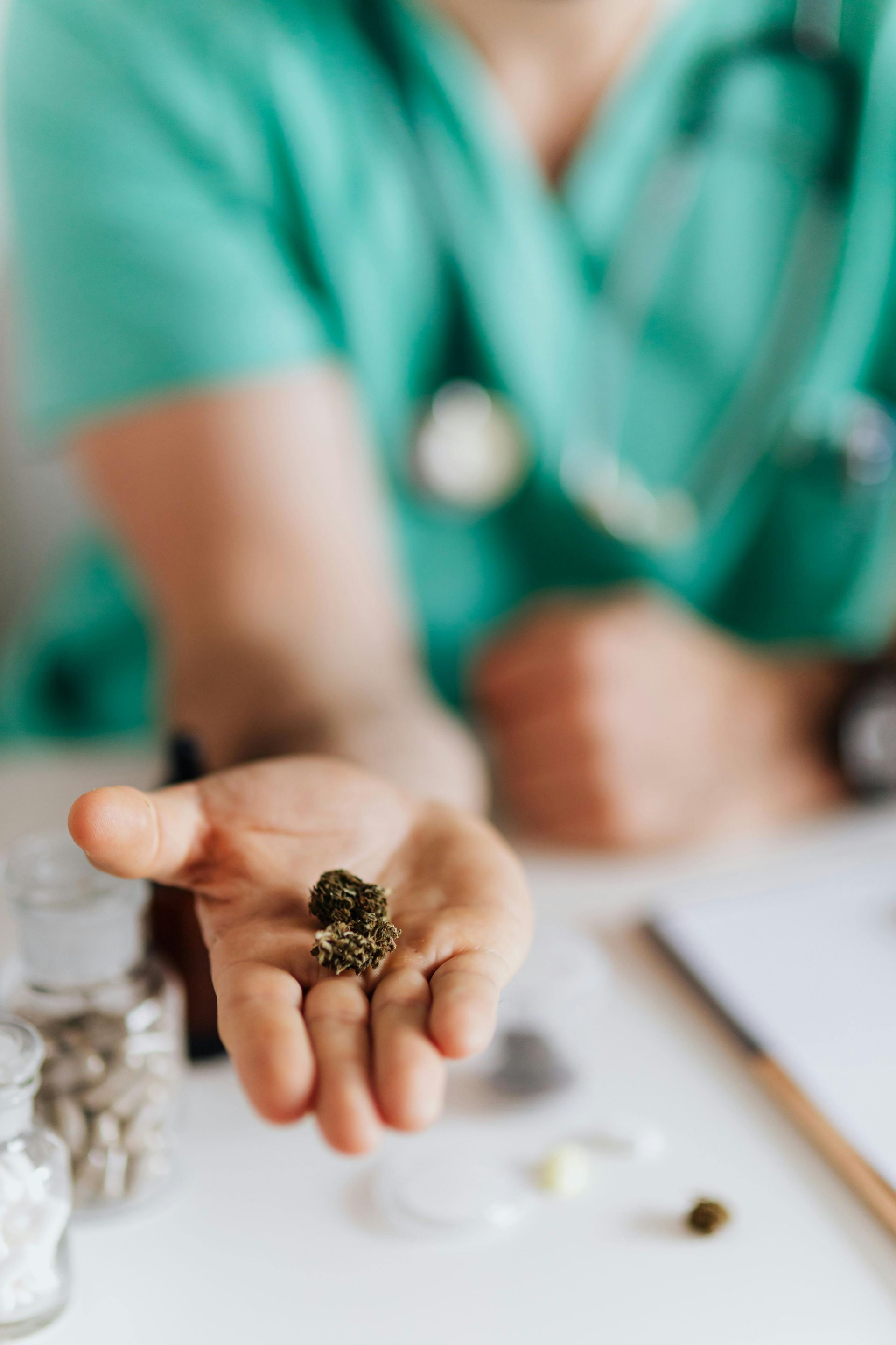 Médico segurando brotos de cannabis na palma da mão. Vestindo uniforme verde, com frascos de remédios sobre a mesa.