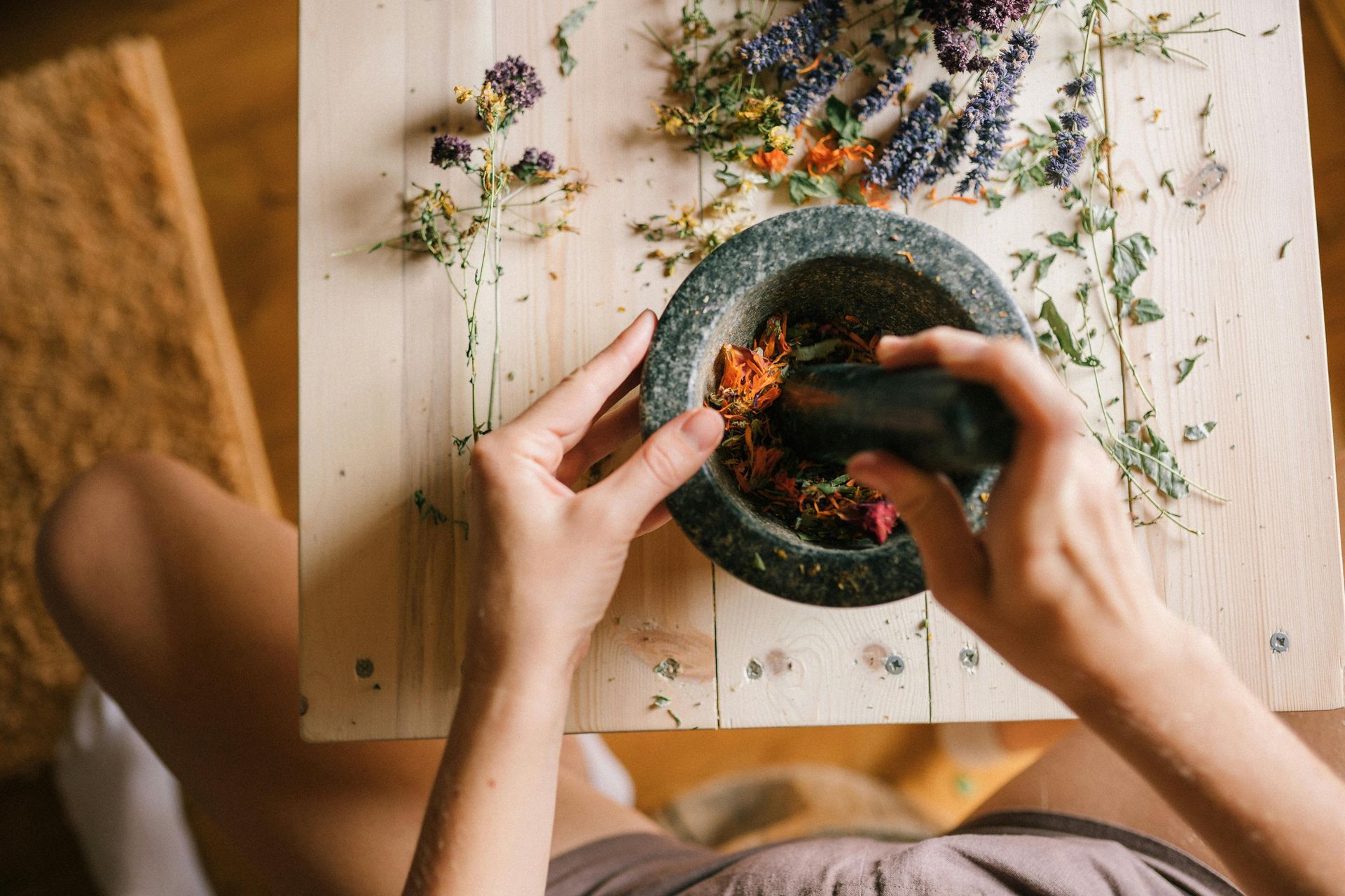 Pessoa moendo ervas em um pilão, cercada por flores secas em uma superfície de madeira.