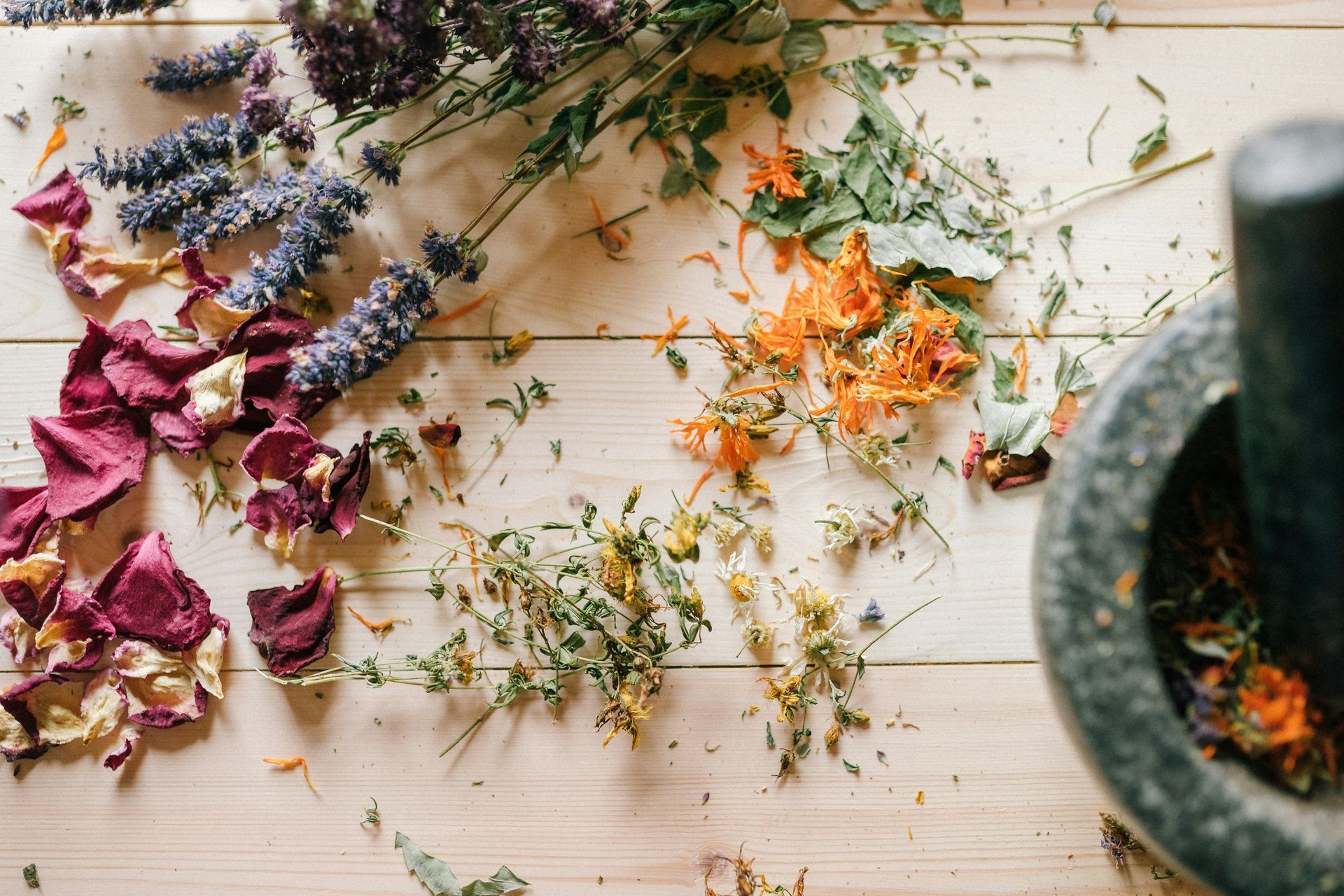 Ervas secas e pétalas de flores espalhadas sobre uma superfície de madeira, com um pilão e um almofariz no lado direito.