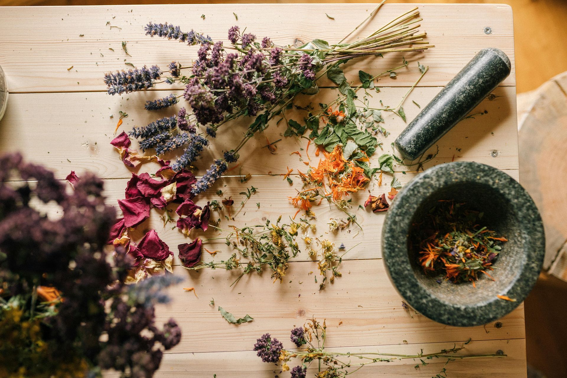 Foto aérea de ervas e flores secas em uma superfície de madeira com um pilão e um almofariz, sugerindo preparação de ervas.