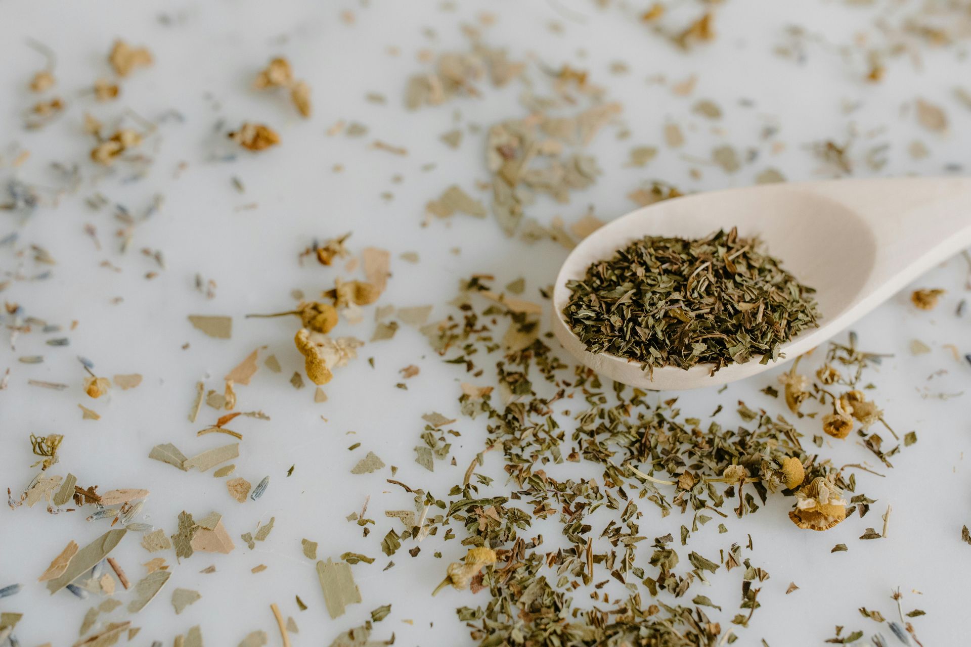 Colher de pau com ervas verdes secas sobre uma superfície branca salpicada com flores e ervas secas.