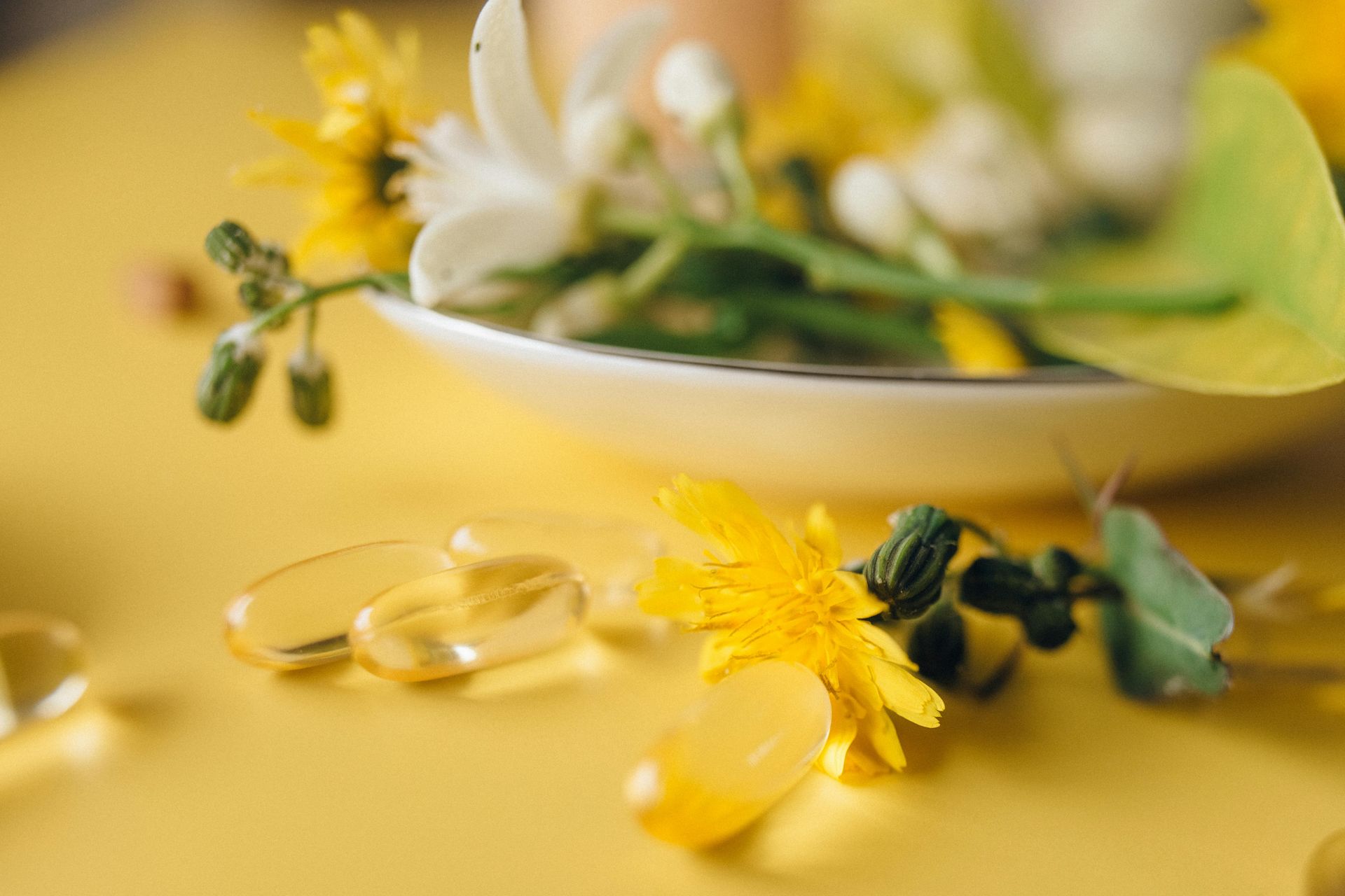 Suplementos amarelos ao lado de um pequeno vaso de flores e folhas em uma superfície amarela.