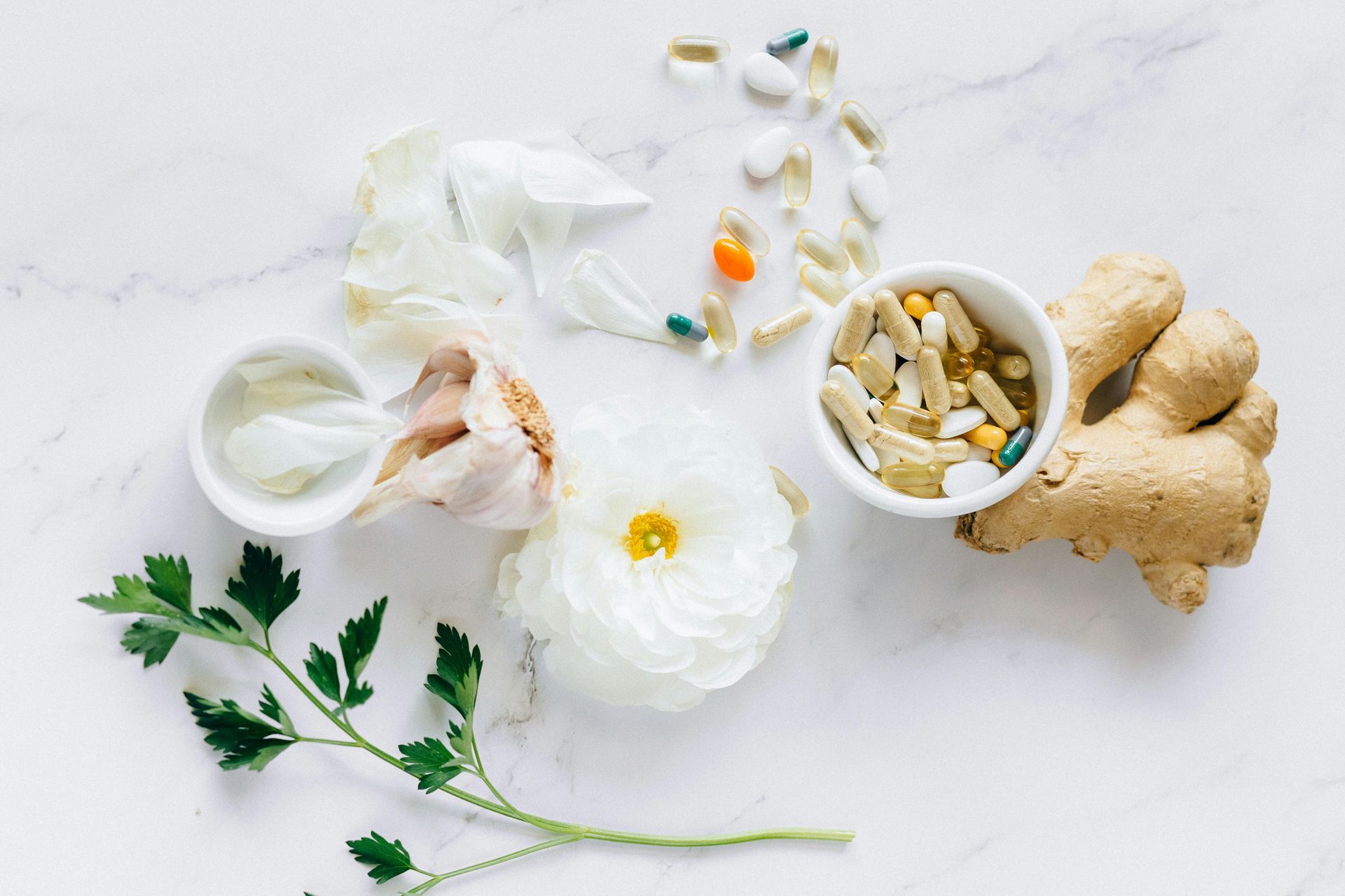 Superfície de mármore branco com alho, salsa, gengibre, pílulas, uma flor branca e uma pequena tigela de suplementos.