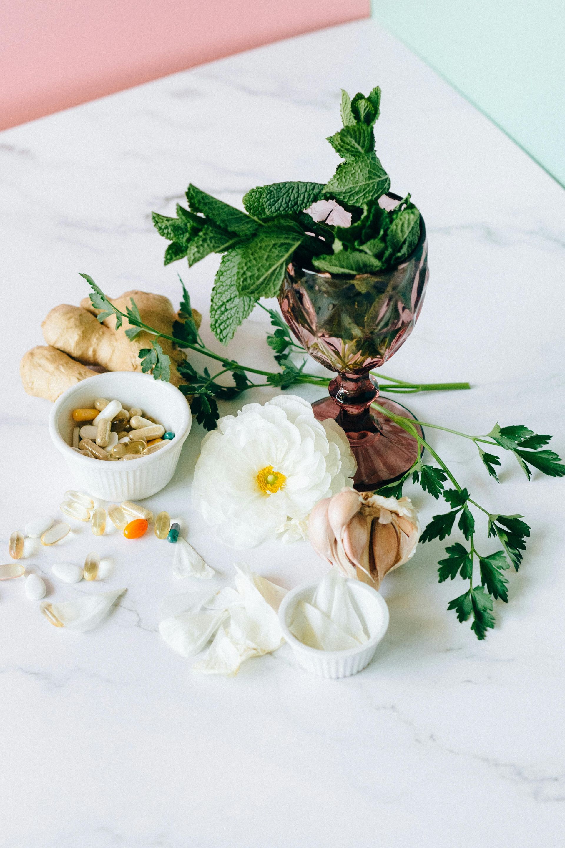 Flor branca, ervas variadas, alho e pílulas dispostas sobre uma superfície de mármore, com fundos rosa e verde.