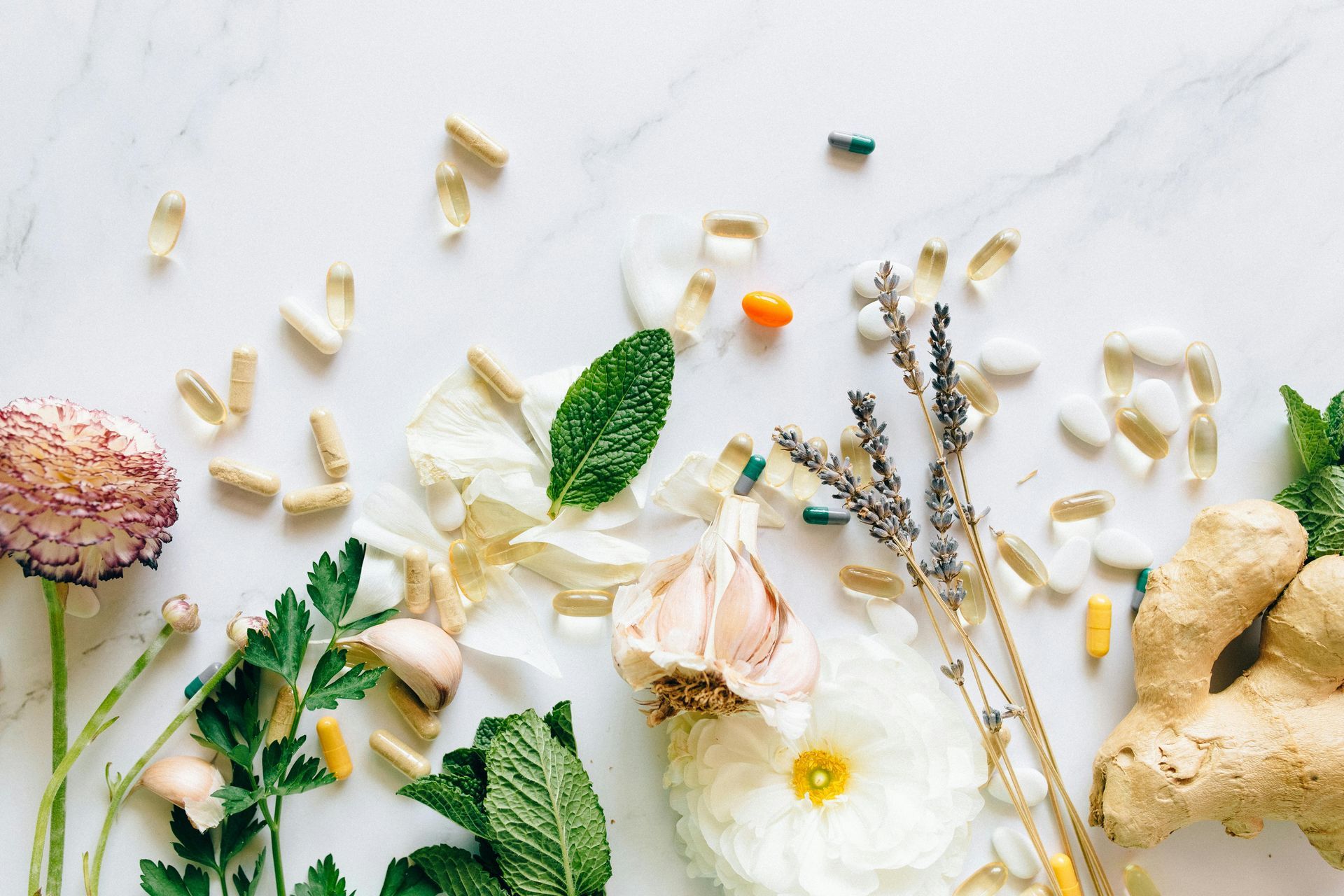 Pílulas e suplementos espalhados com flores, ervas e gengibre em uma superfície de mármore branco.
