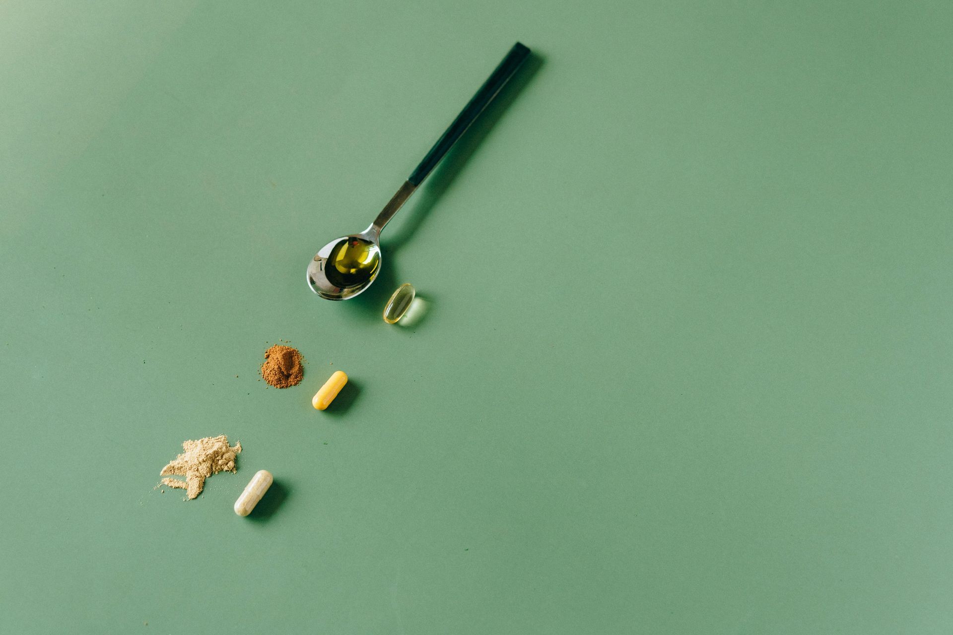 Suplementos e colher sobre uma superfície verde. Diversos comprimidos e uma colher de líquido estão espalhados.