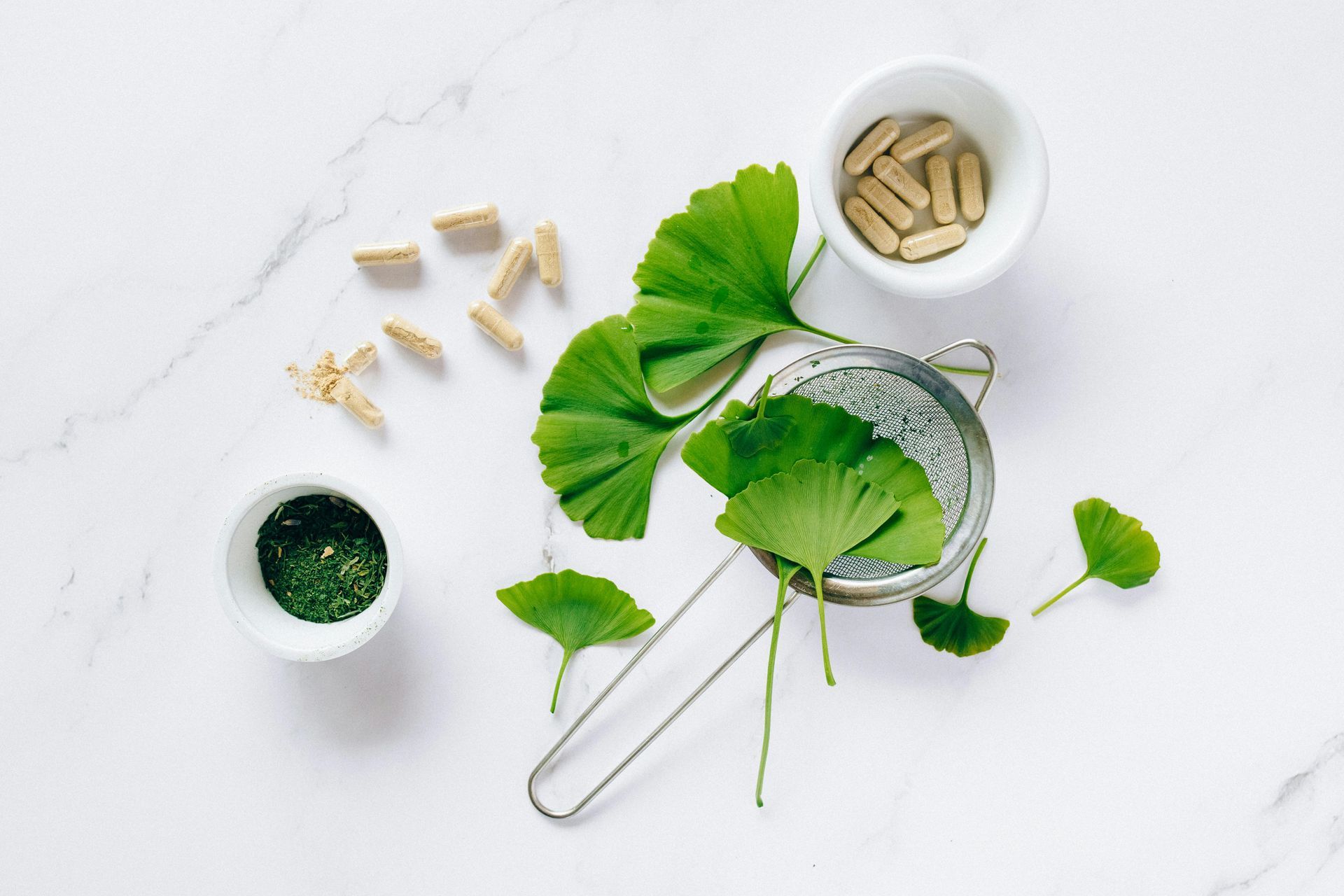 Folhas de ginkgo, suplementos de ervas e um coador dispostos sobre uma superfície de mármore branco.