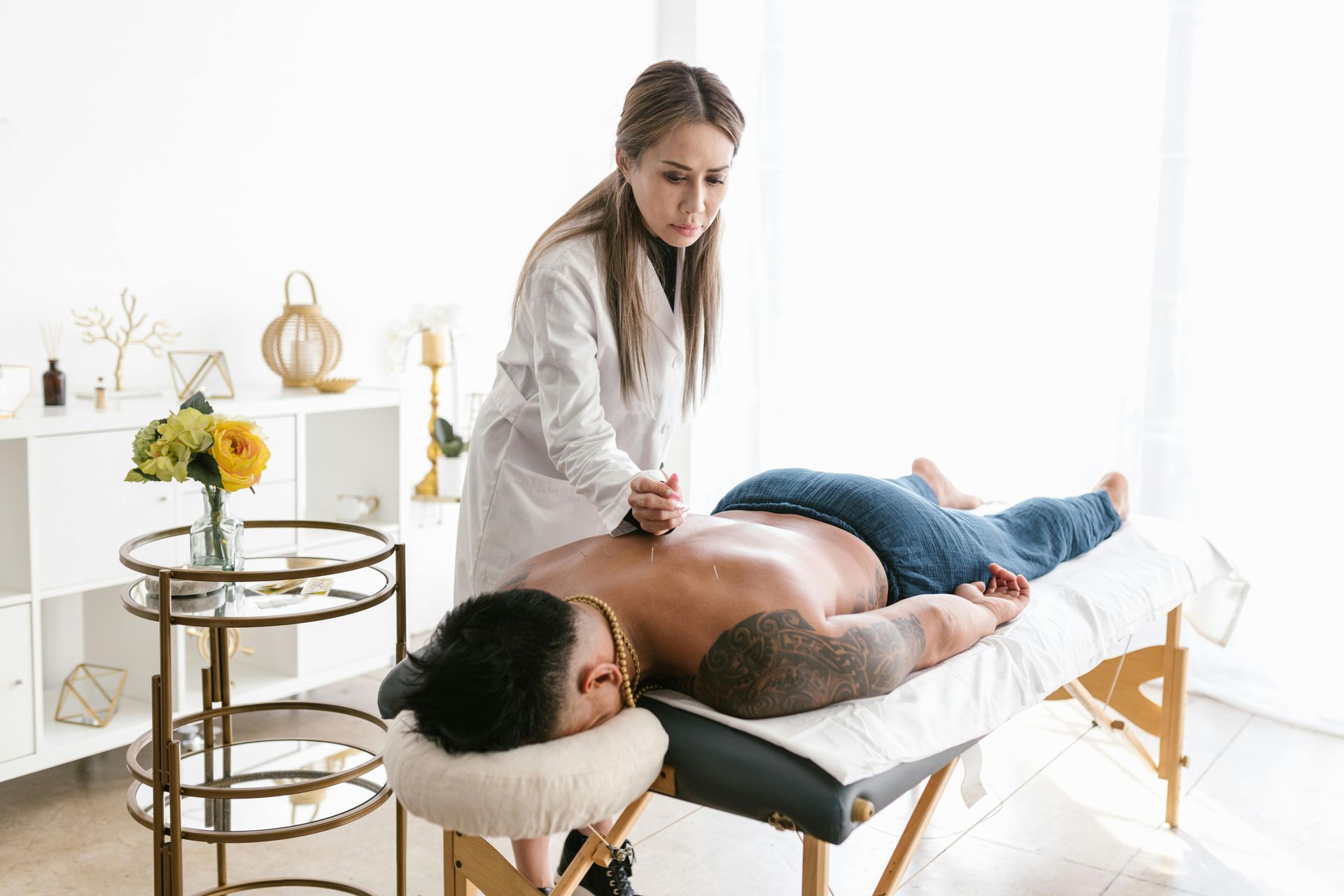 Uma massagista de jaleco branco massageia as costas de um homem. Eles estão em uma sala iluminada com uma mesa de massagem.