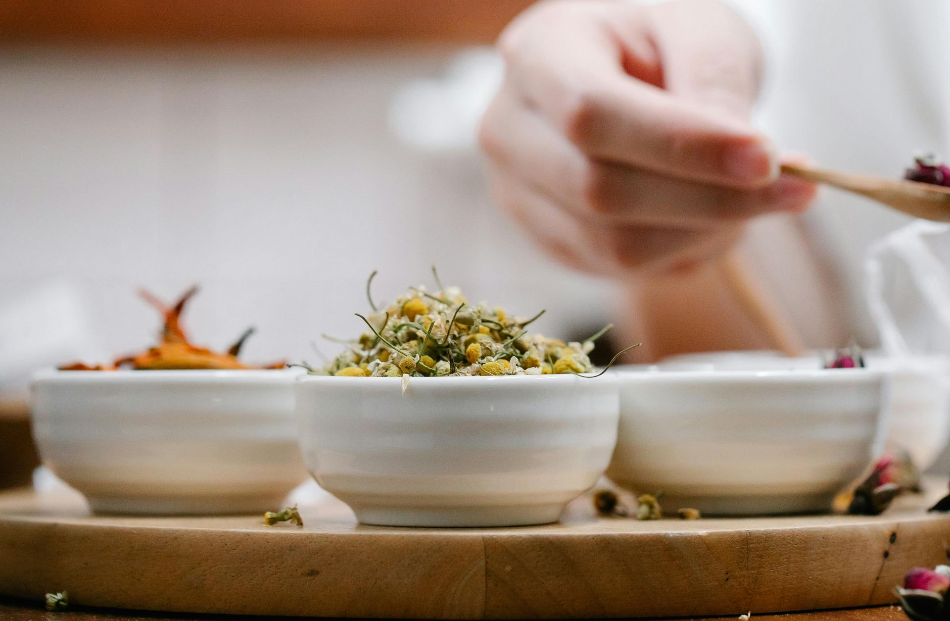 Pessoa usando uma pequena colher de pau para adicionar botões de rosa secos de uma tigela branca ao chá.