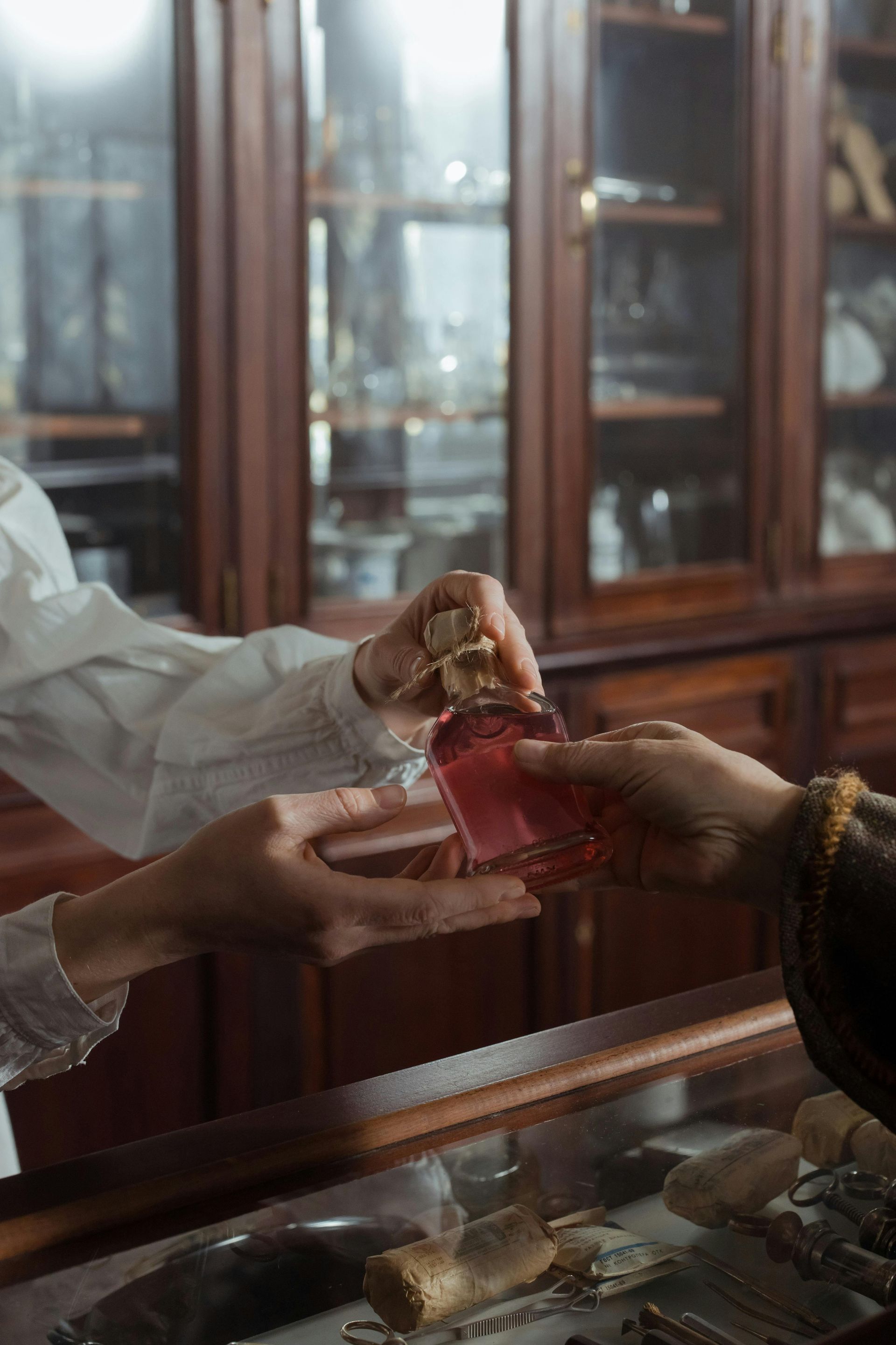 Duas mãos trocando um frasco de perfume rosa sobre uma vitrine de vidro em uma loja com painéis de madeira.