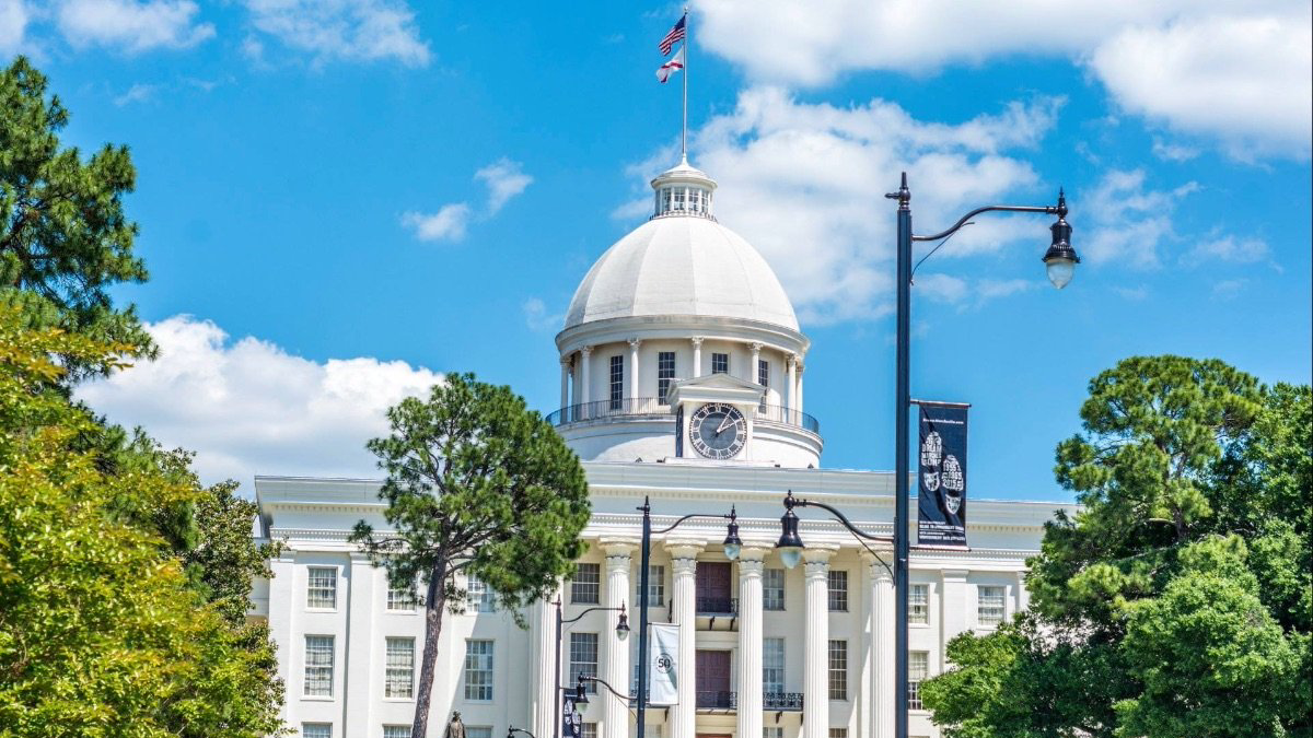 The Alabama State Capitol building featuring a white dome and columns under a blue sky with trees in the foreground.