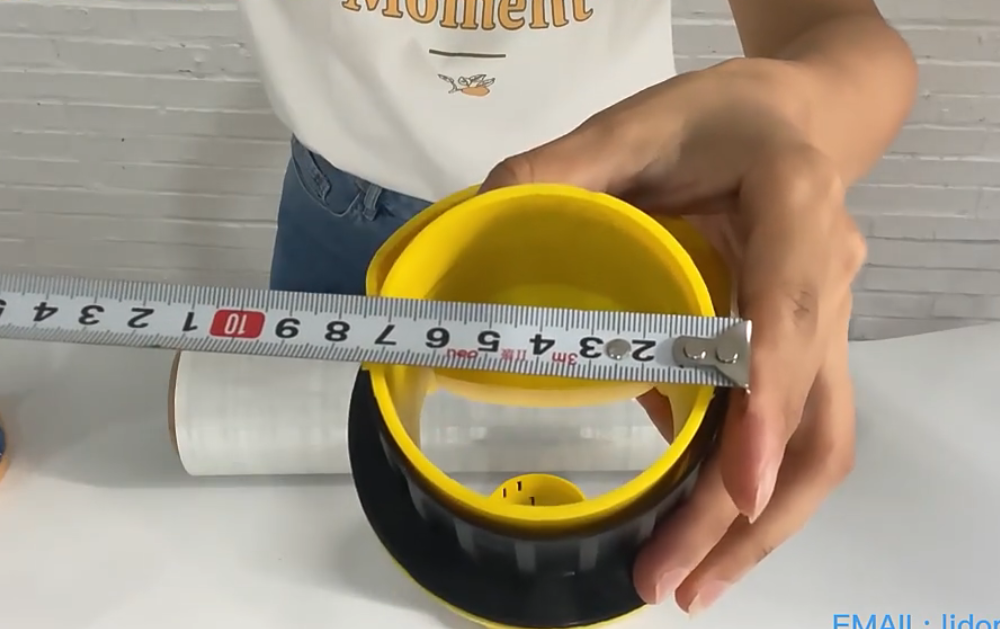 Una persona sostiene un tubo grande de plástico transparente con extremos amarillos sobre una mesa blanca, con cinta adhesiva cerca.