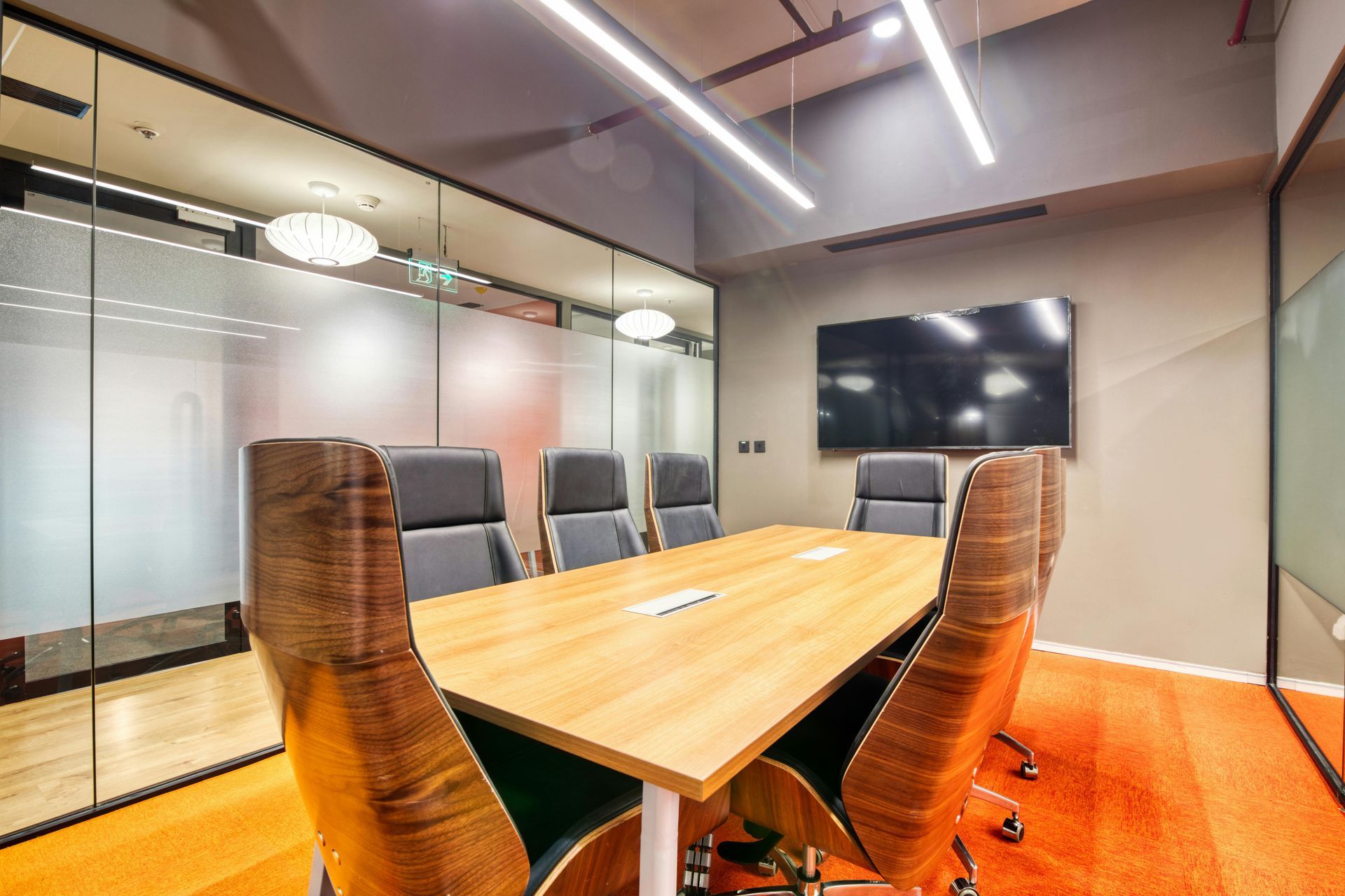 Moderna sala de conferencias con mesa de madera, sillas, paredes de cristal y televisor montado en la pared.