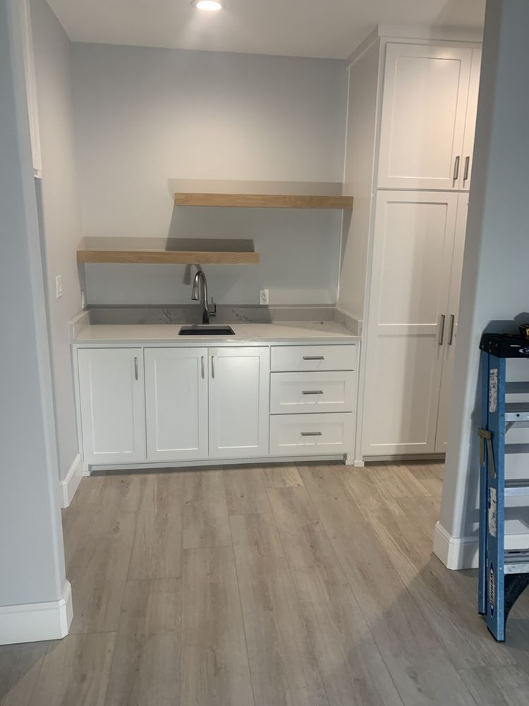 Small white kitchenette with sink, cabinets, wood shelf, and light wood flooring.