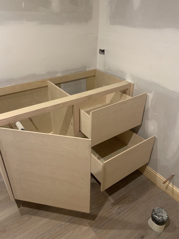 Unfinished corner cabinet with open drawers in a room with bare walls and flooring.