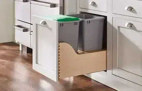 Pull-out kitchen cabinet drawer with a gray trash bin and green lid beside white cabinets and wood floor