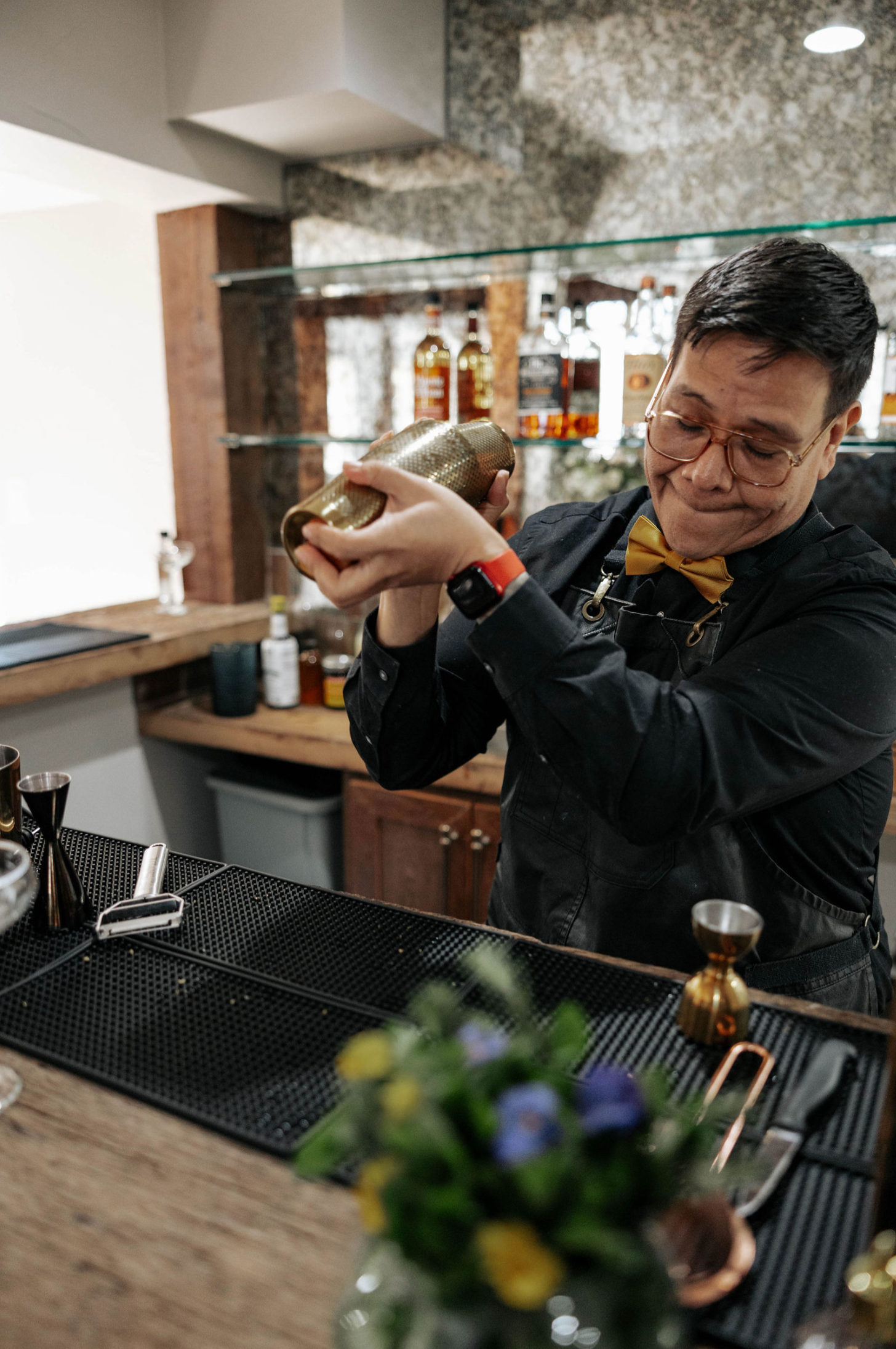 Bartender shaking a cocktail at a bar, wearing a yellow bow tie and vest, with a floral arrangement in the foreground.