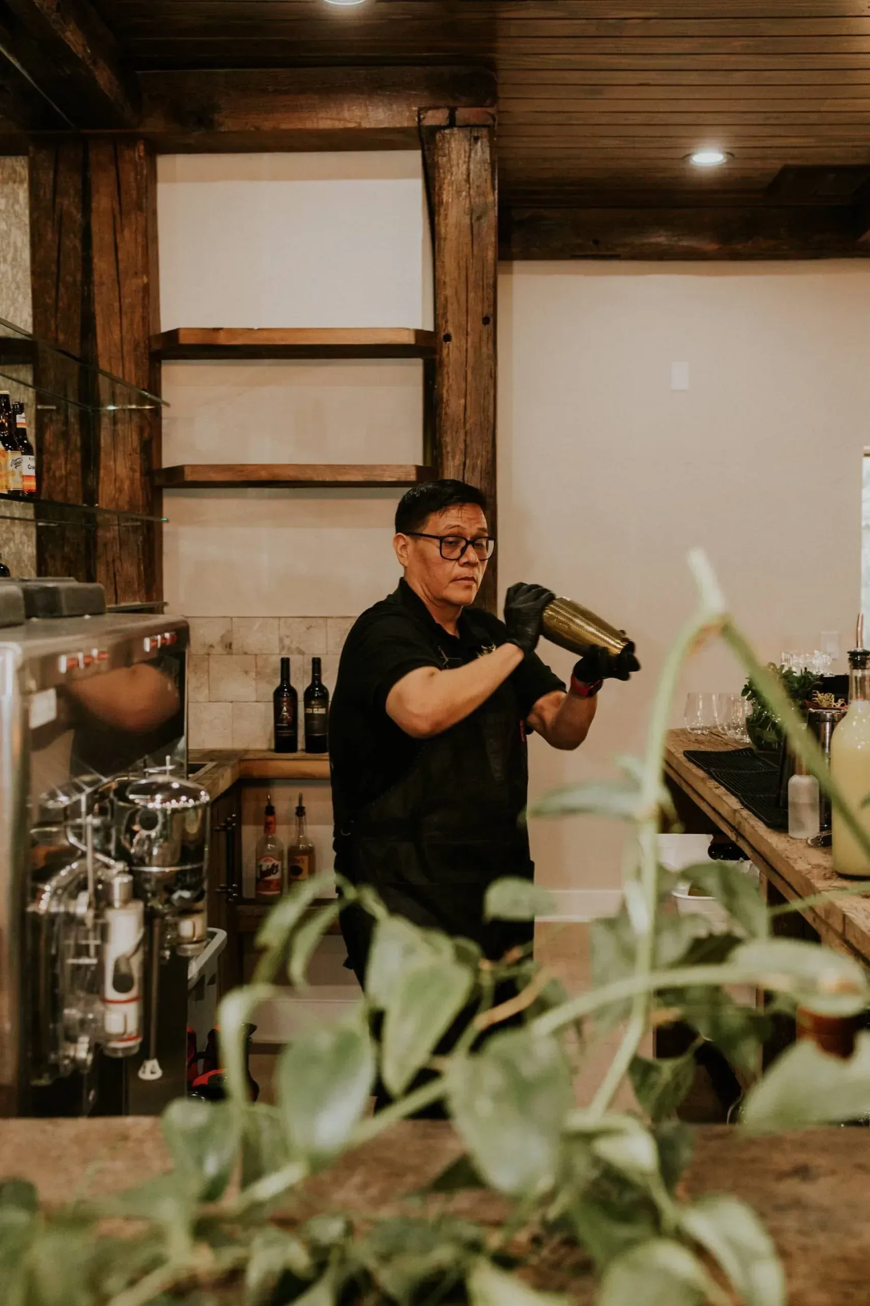Bartender shaking a cocktail at a bar. Wooden shelves and decorative plants are in the background.