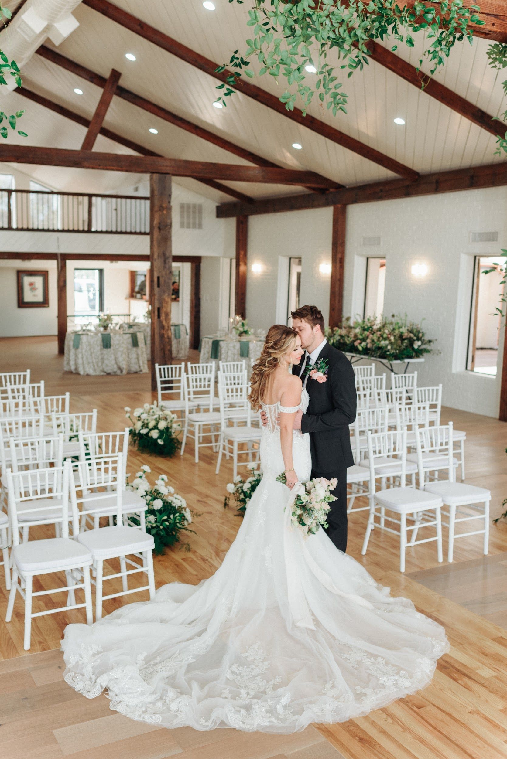 Bride and groom embrace in a wedding ceremony venue. White chairs line an aisle, florals and greenery decorate the space.