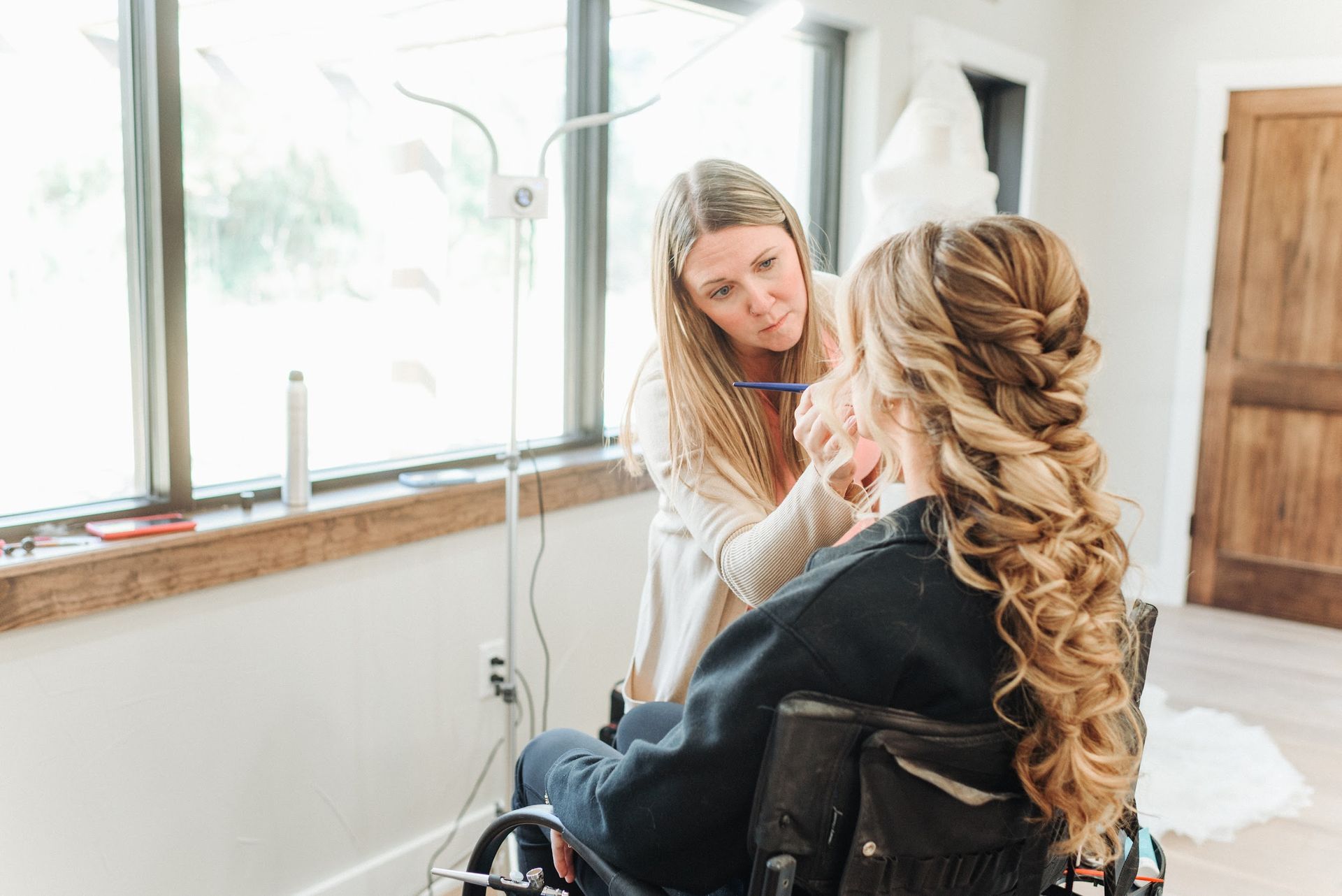 Makeup artist applies makeup to a person in a wheelchair; natural light, braided hairstyle.