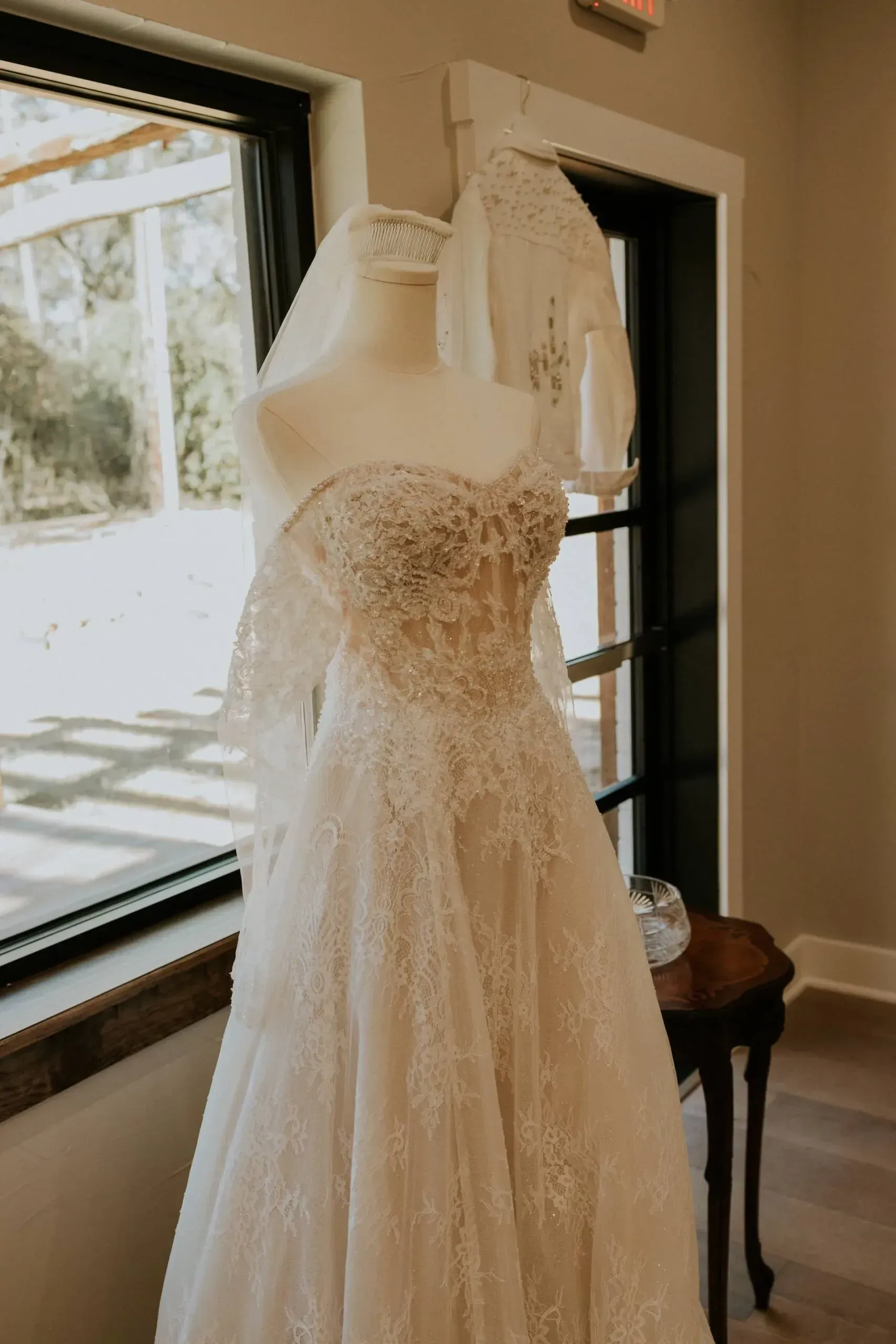 Wedding dress on a mannequin next to a window; veil, lace, and beading details.