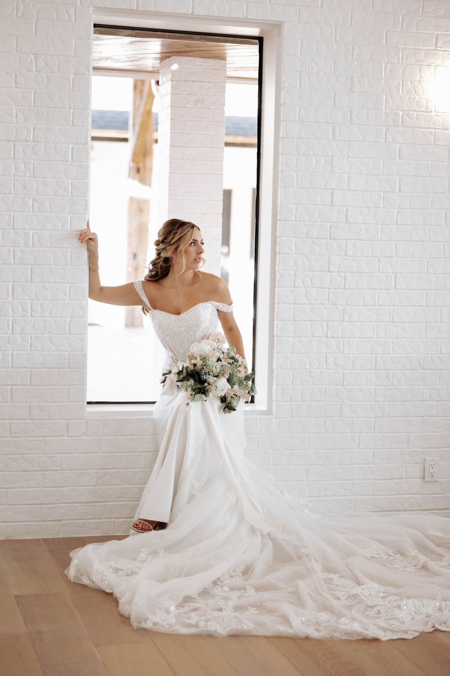 Bride in white wedding dress, leaning in window, holding bouquet. Brick wall background.