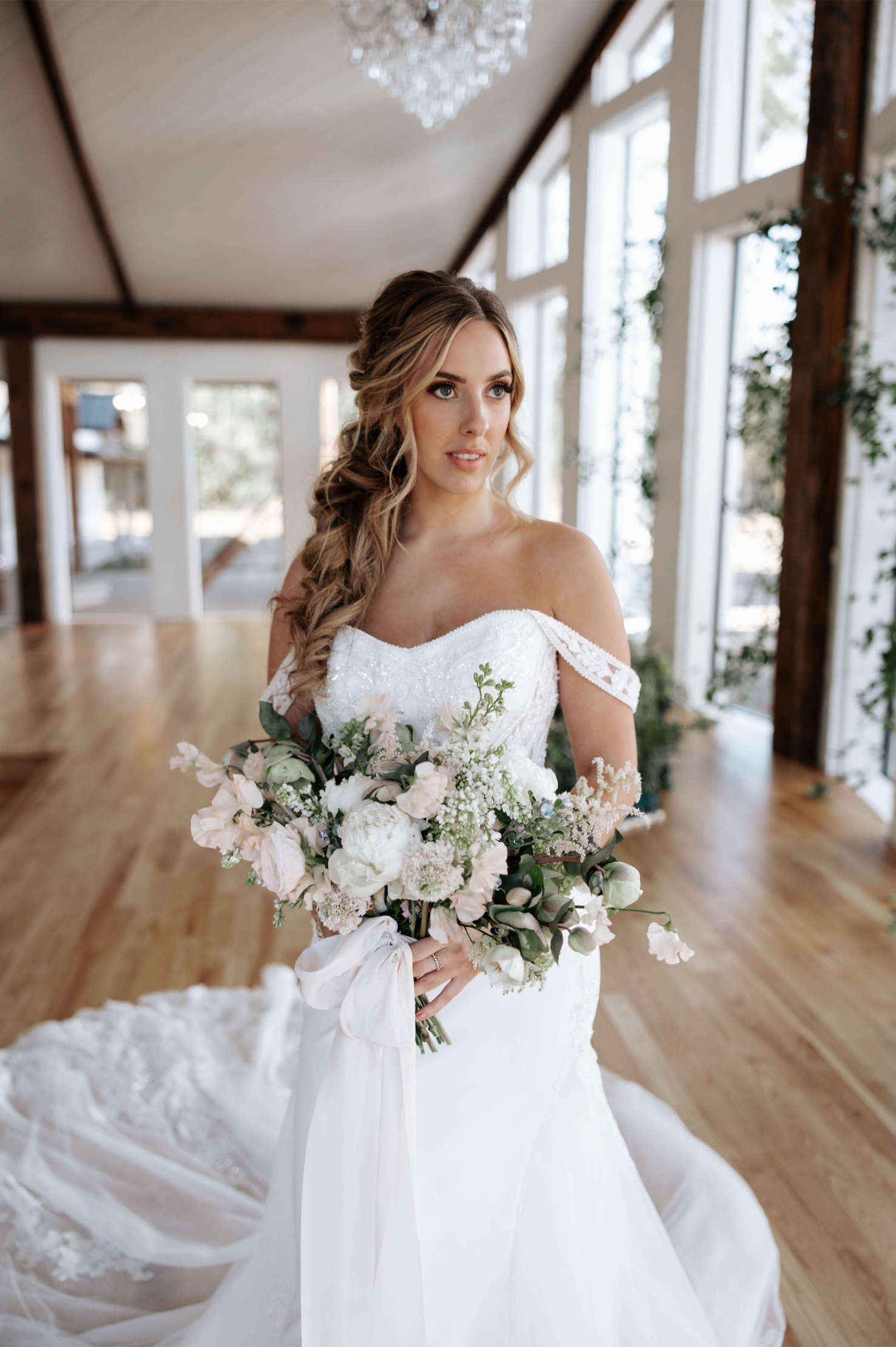 Bride in off-the-shoulder gown, holding a bouquet, in a bright, airy room with large windows.