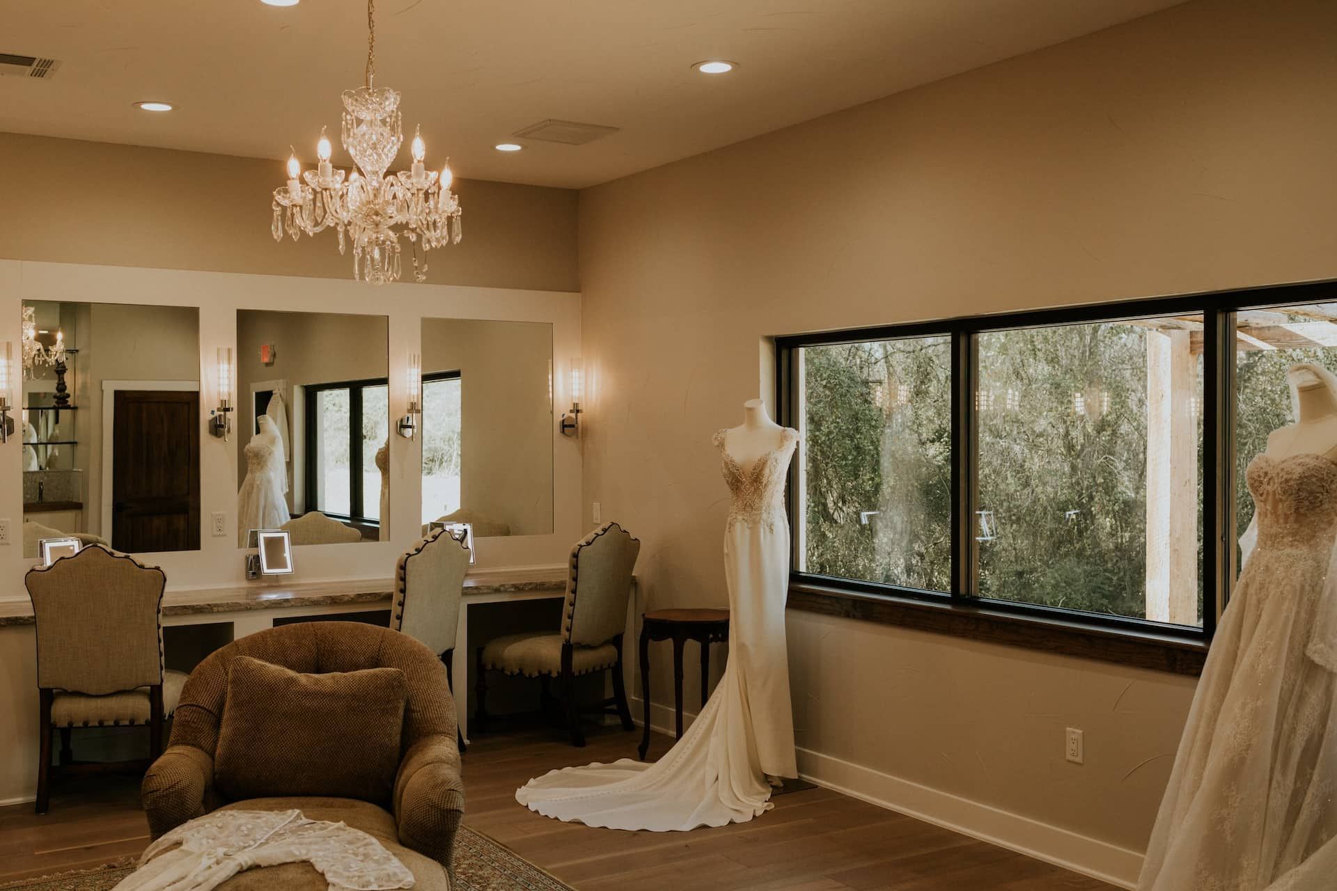 Bridal suite with mirrors, gowns, and a chandelier; a window offers a view of greenery.
