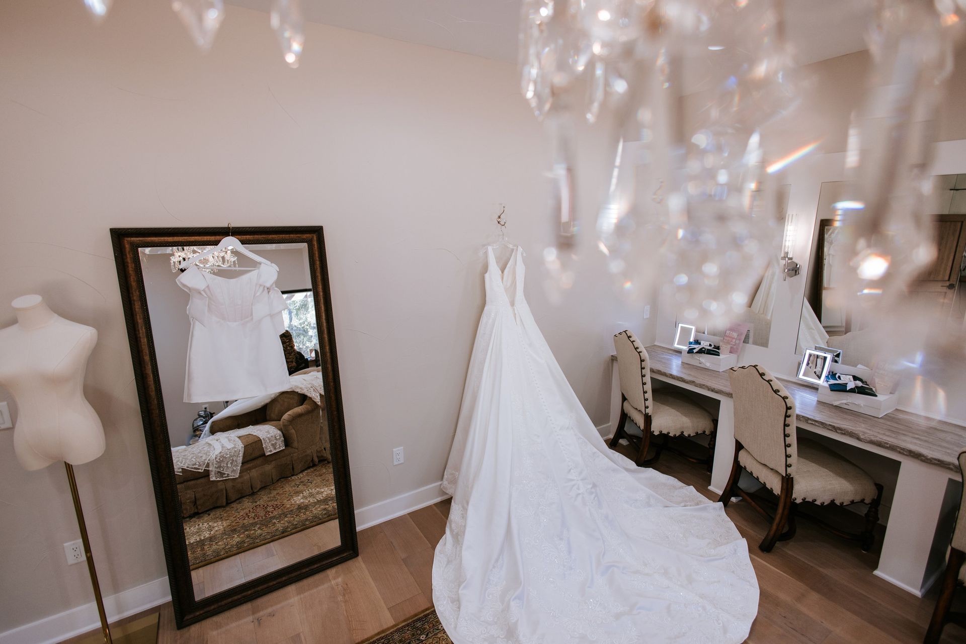 A wedding dress and bridesmaid dress hang in a room with a mirror and vanity.