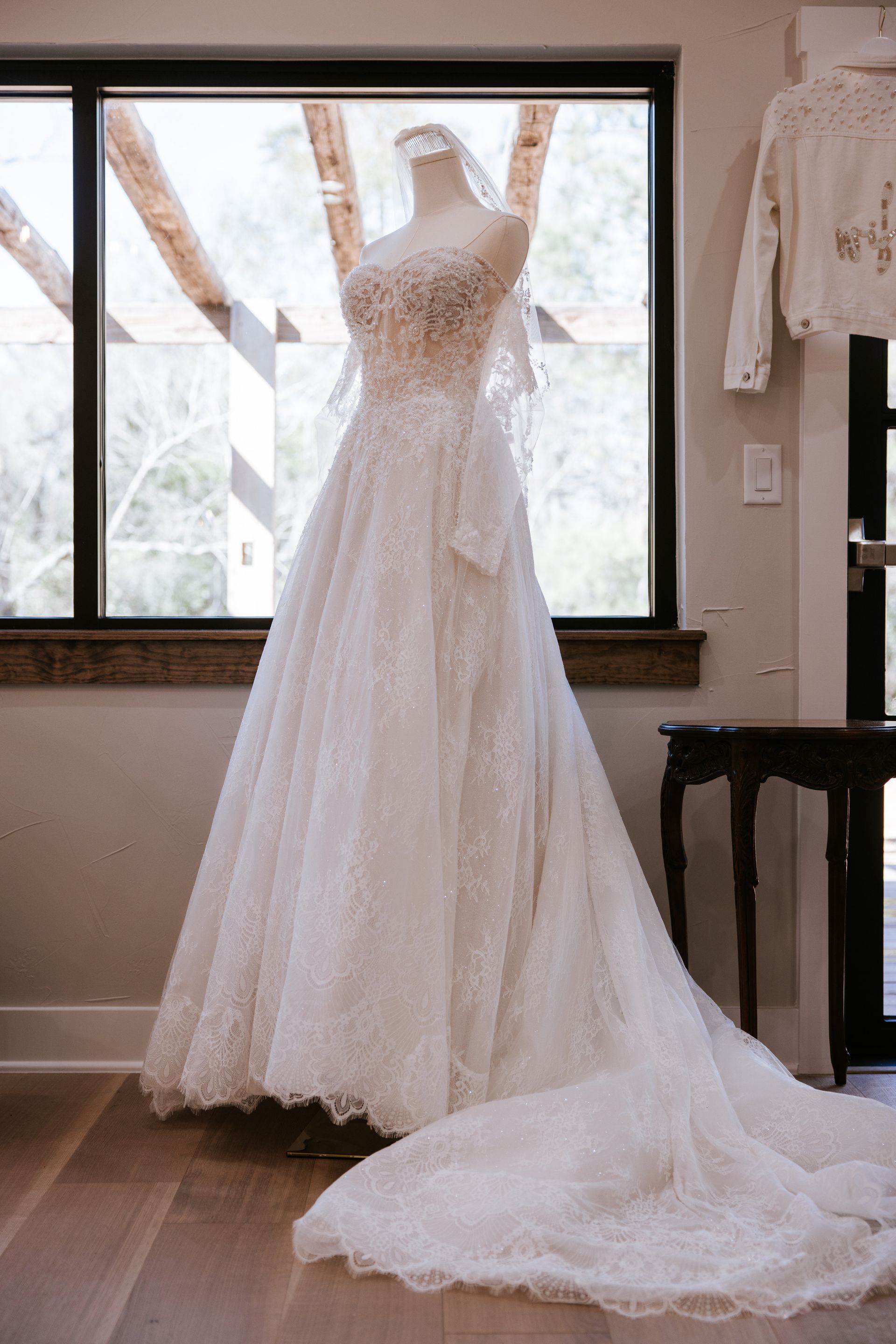 Wedding dress on a mannequin, displayed indoors near a window. The gown is white with intricate details, train extended on the floor.