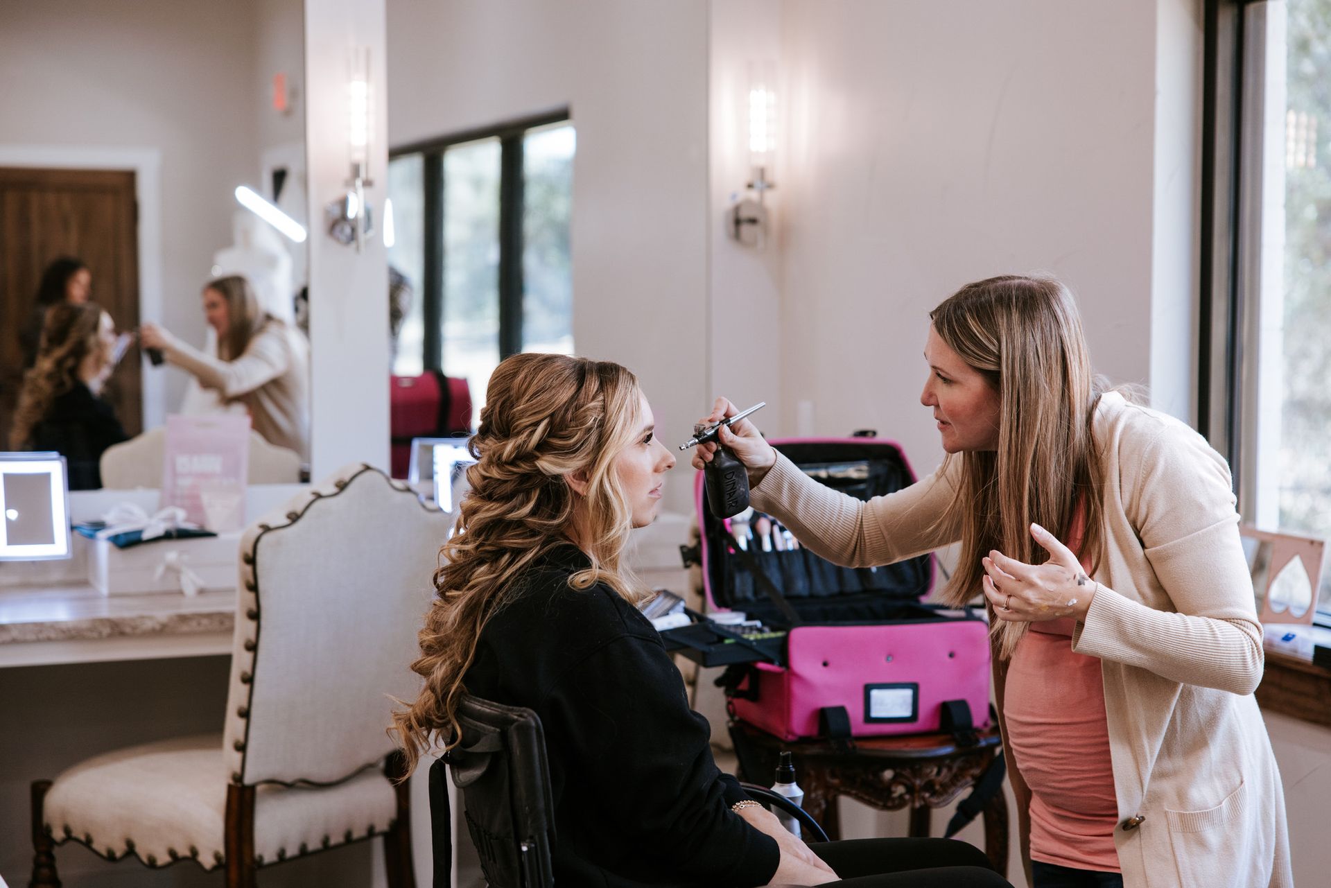 Makeup artist applying makeup to a client in a well-lit room with mirrors and a pink makeup case.