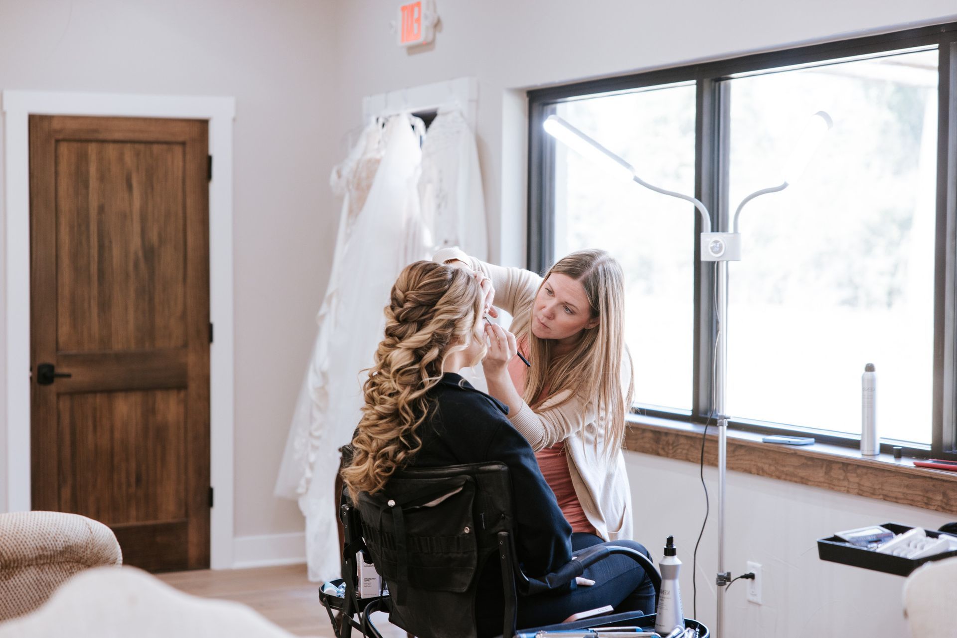 Woman having makeup applied in a room with a dress hanging in the background, near a window.