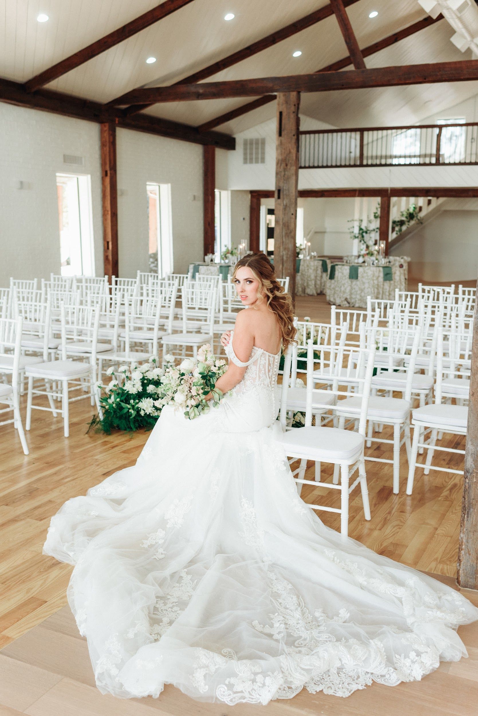 Bride in a white gown holding bouquet, posing in a wedding venue with white chairs and floral decor.