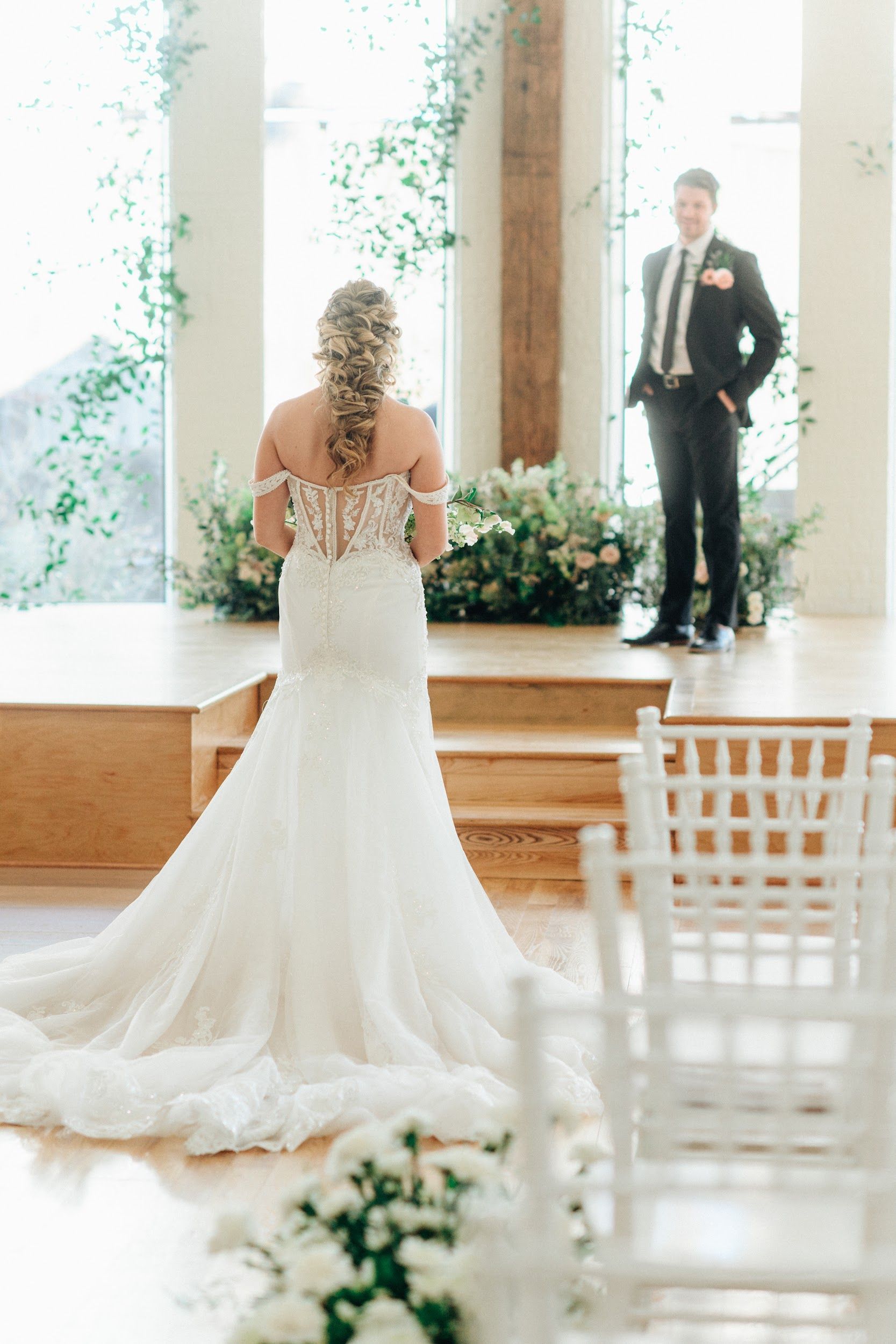 Bride in lace gown walks toward groom at altar, floral arch, sunny interior.