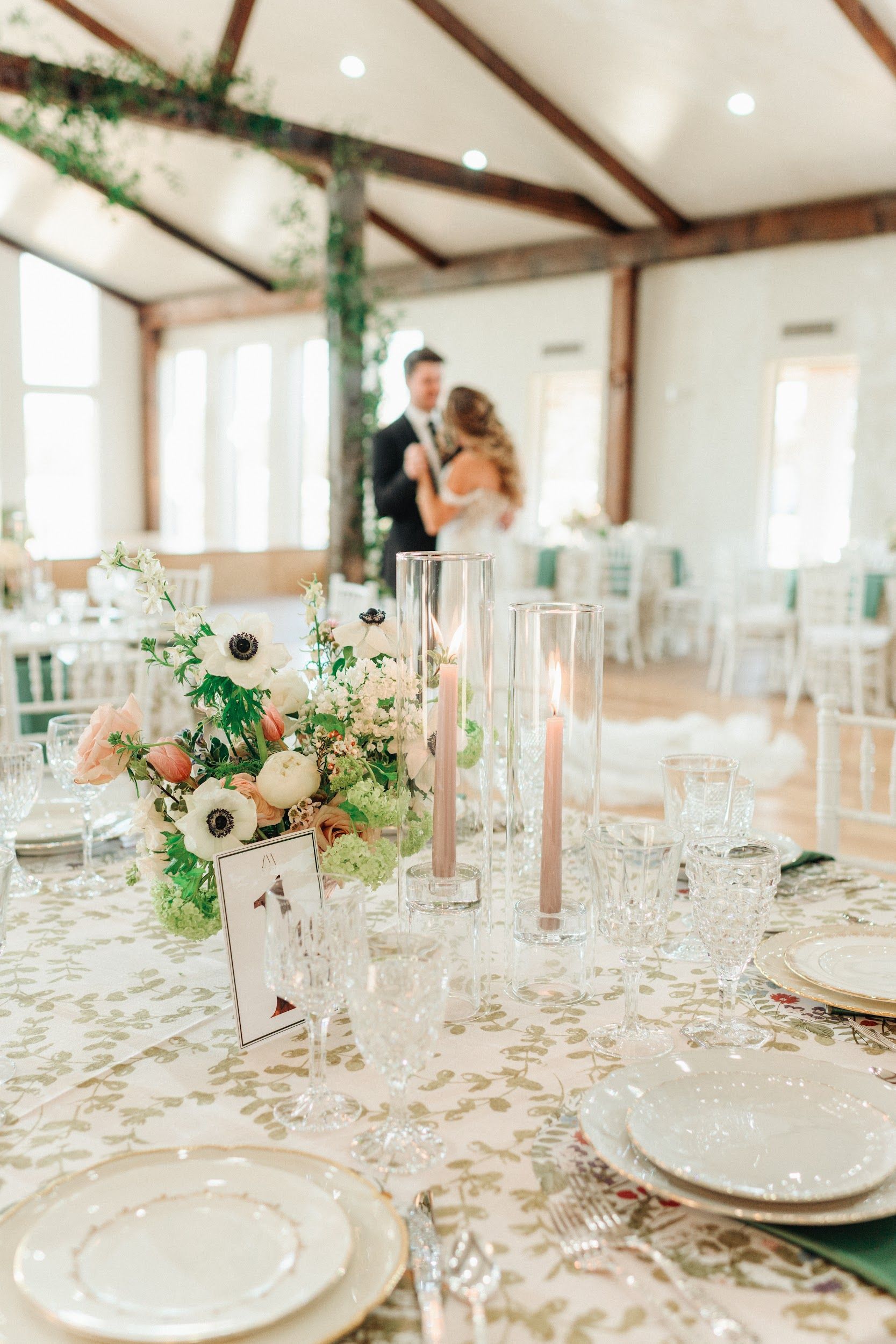 Wedding reception table setting with floral arrangement and couple dancing in the background.