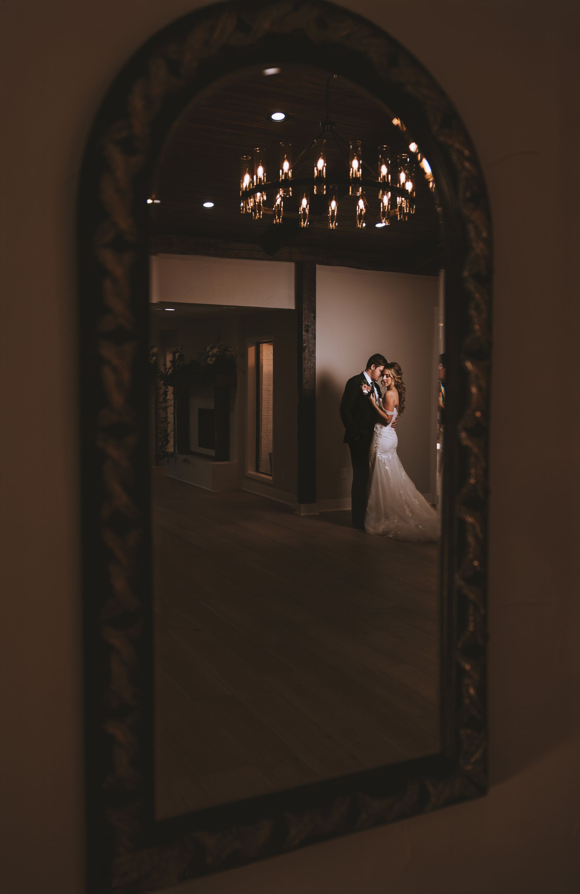 Mirror reflection of a couple in wedding attire embracing indoors. Antique mirror, soft lighting.