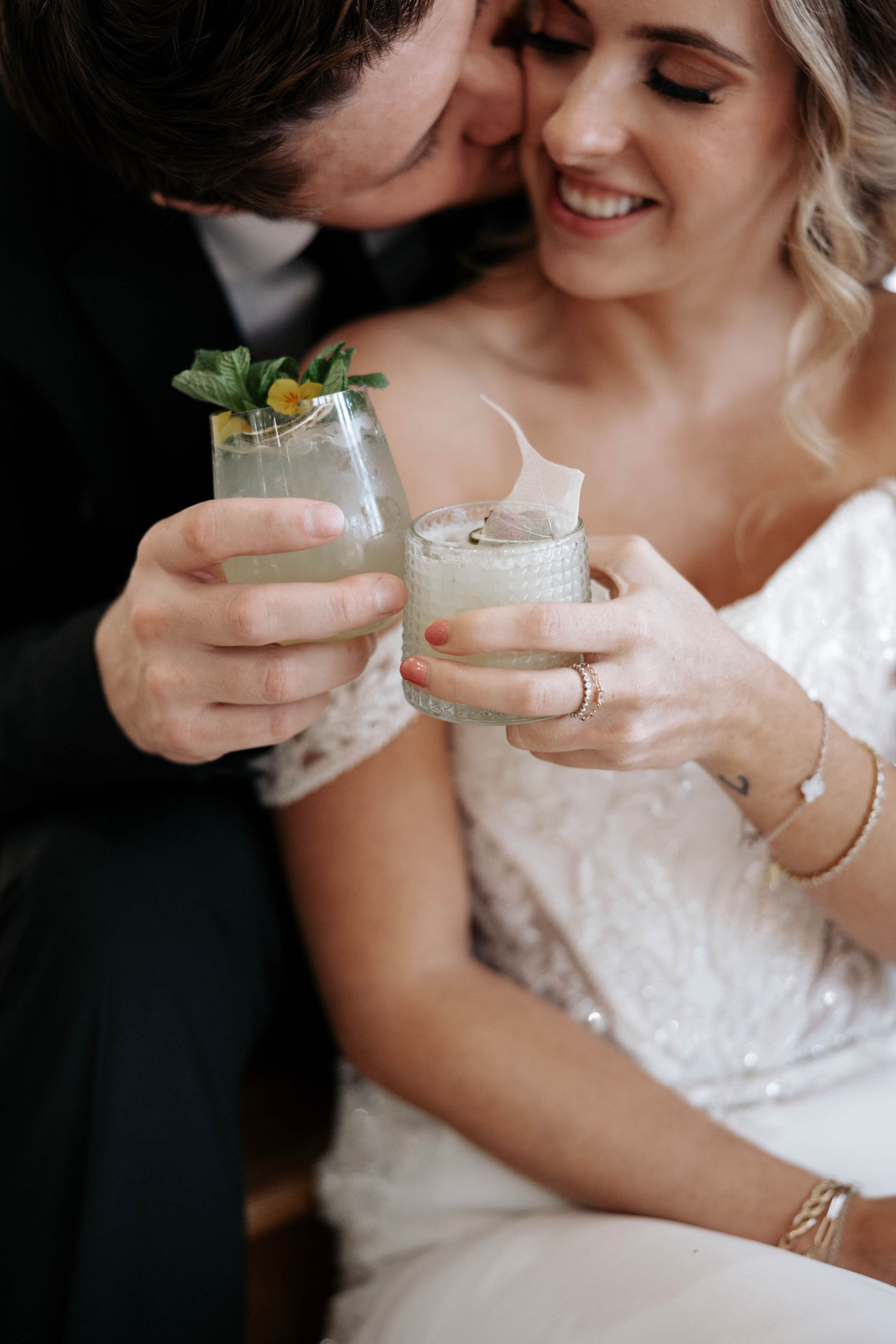 Couple toasting cocktails, man kisses woman's cheek, wearing wedding attire; cocktails garnished with mint and flowers.