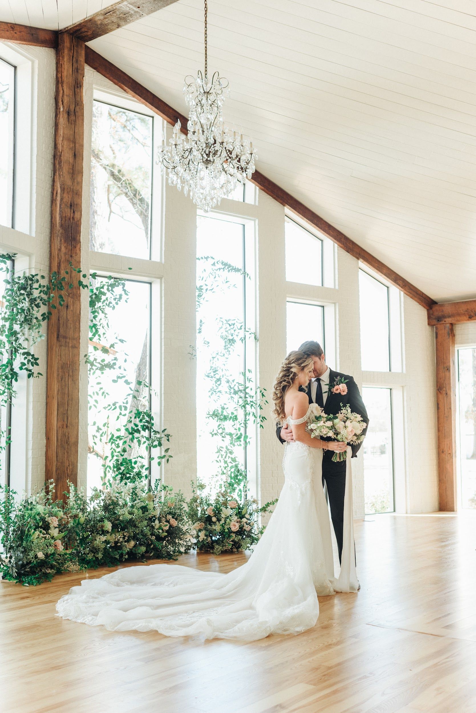 Bride and groom embrace in a light-filled room with large windows, decorated with greenery and a chandelier.