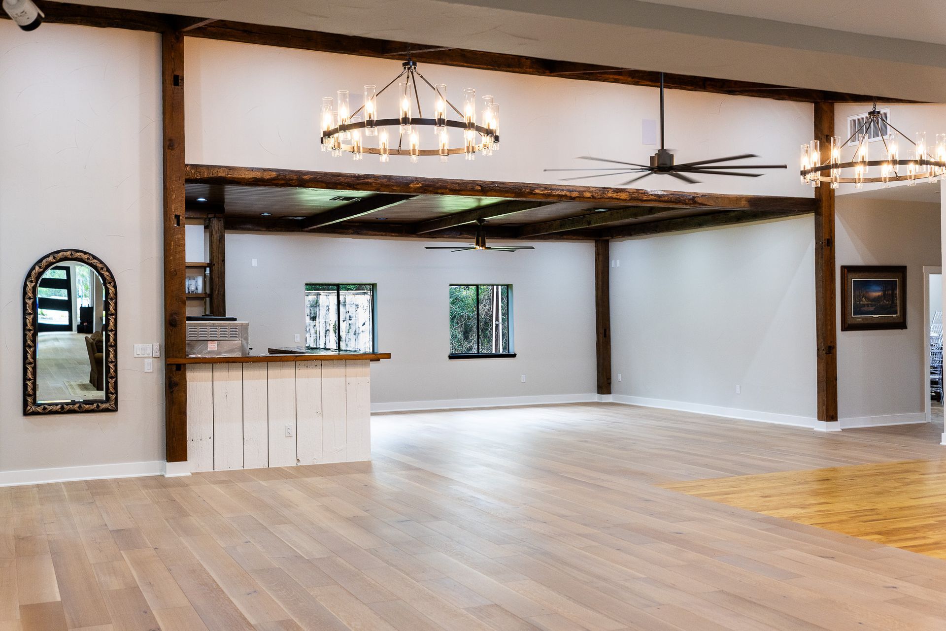 Large, empty room with wooden beams and a dance floor, chandeliers, a bar, and mirrors.