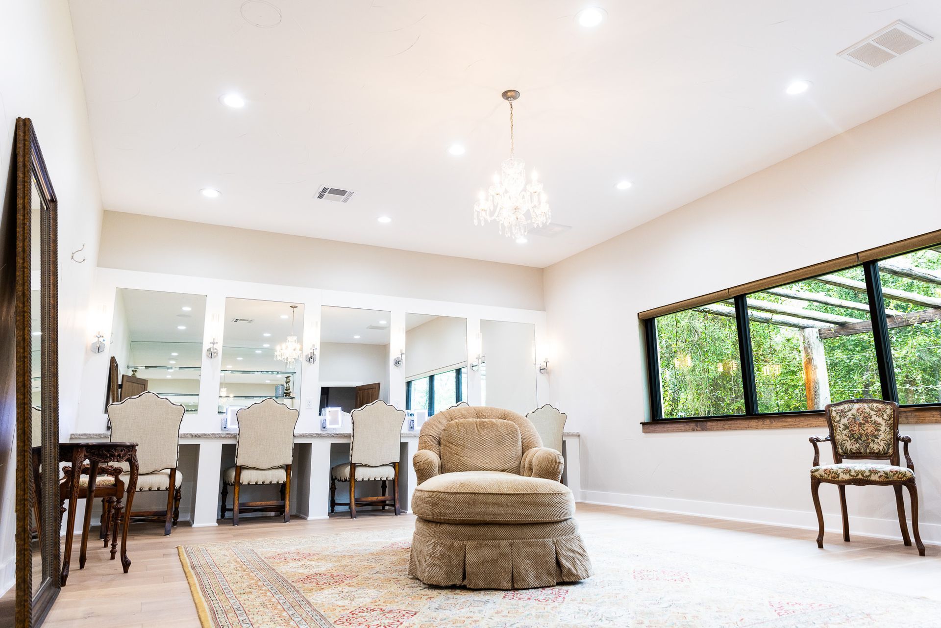Spacious bridal suite with vanity mirrors, chairs, chandelier, large window overlooking greenery.