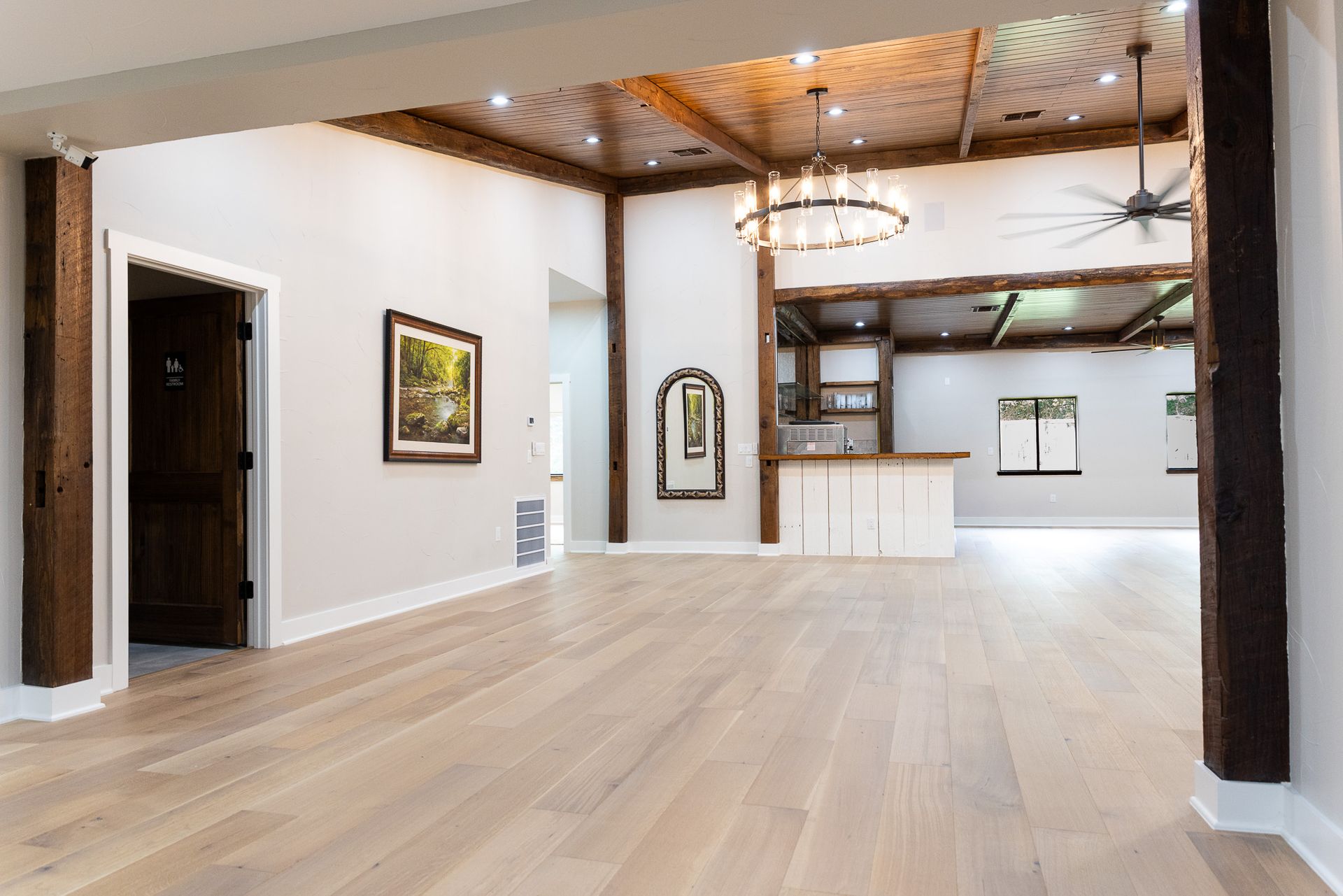 Empty room with light wood floors, white walls, and dark wood trim, leading to a bar area with chandelier.