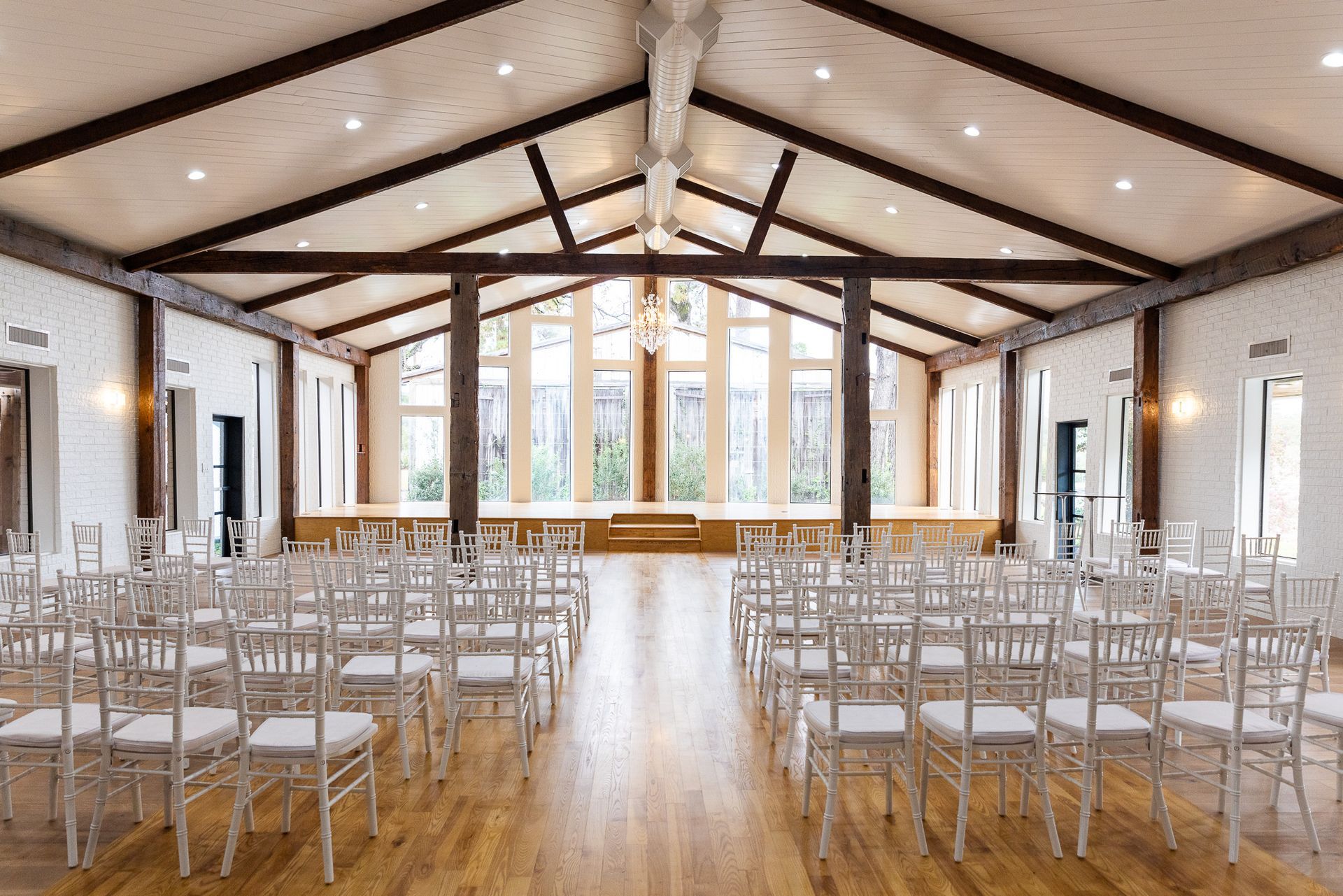 Rows of white chairs face a stage in a wooden-beamed wedding venue with large windows.