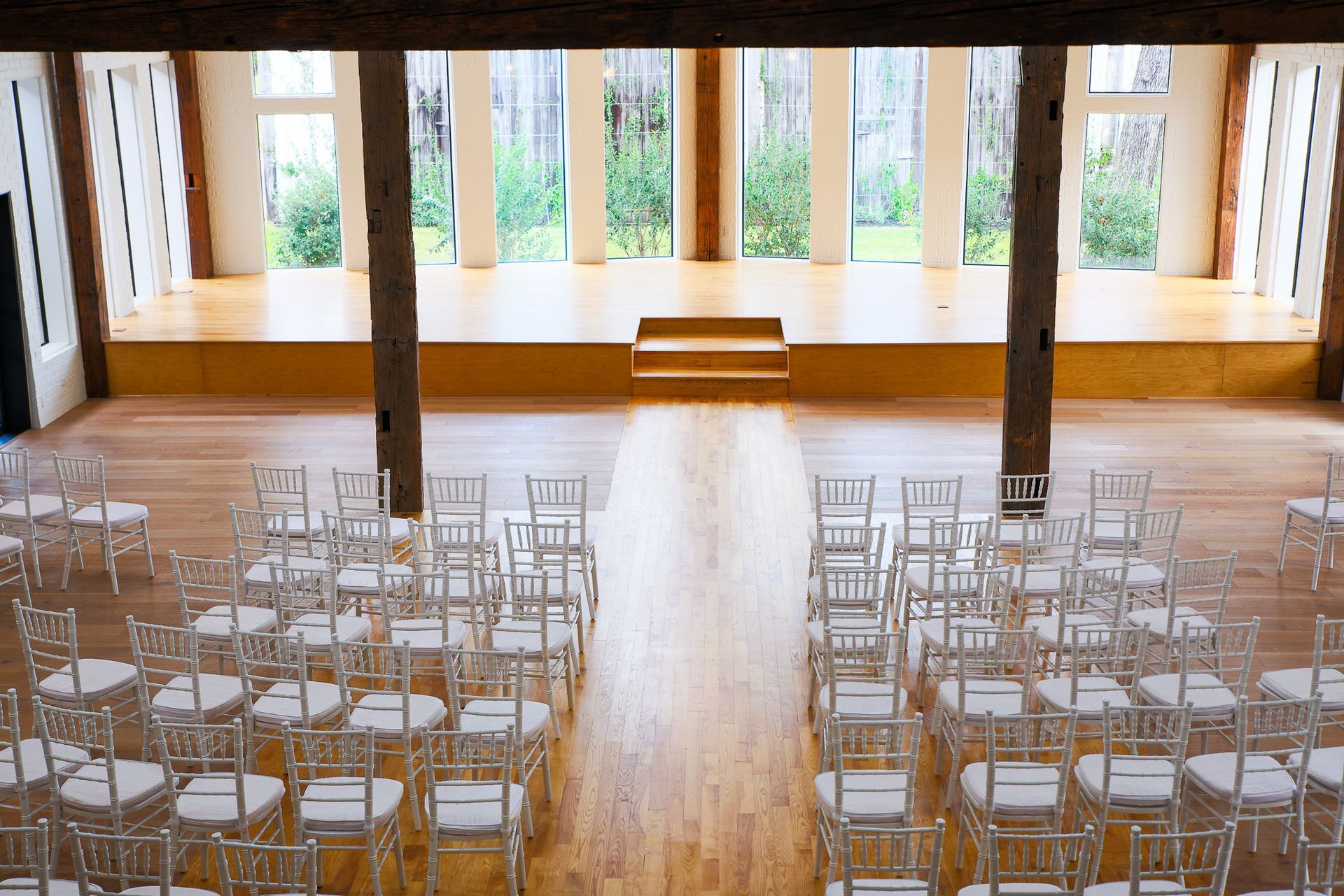 Rows of clear chairs face a stage in a wooden-floored room with large windows, set up for an event.